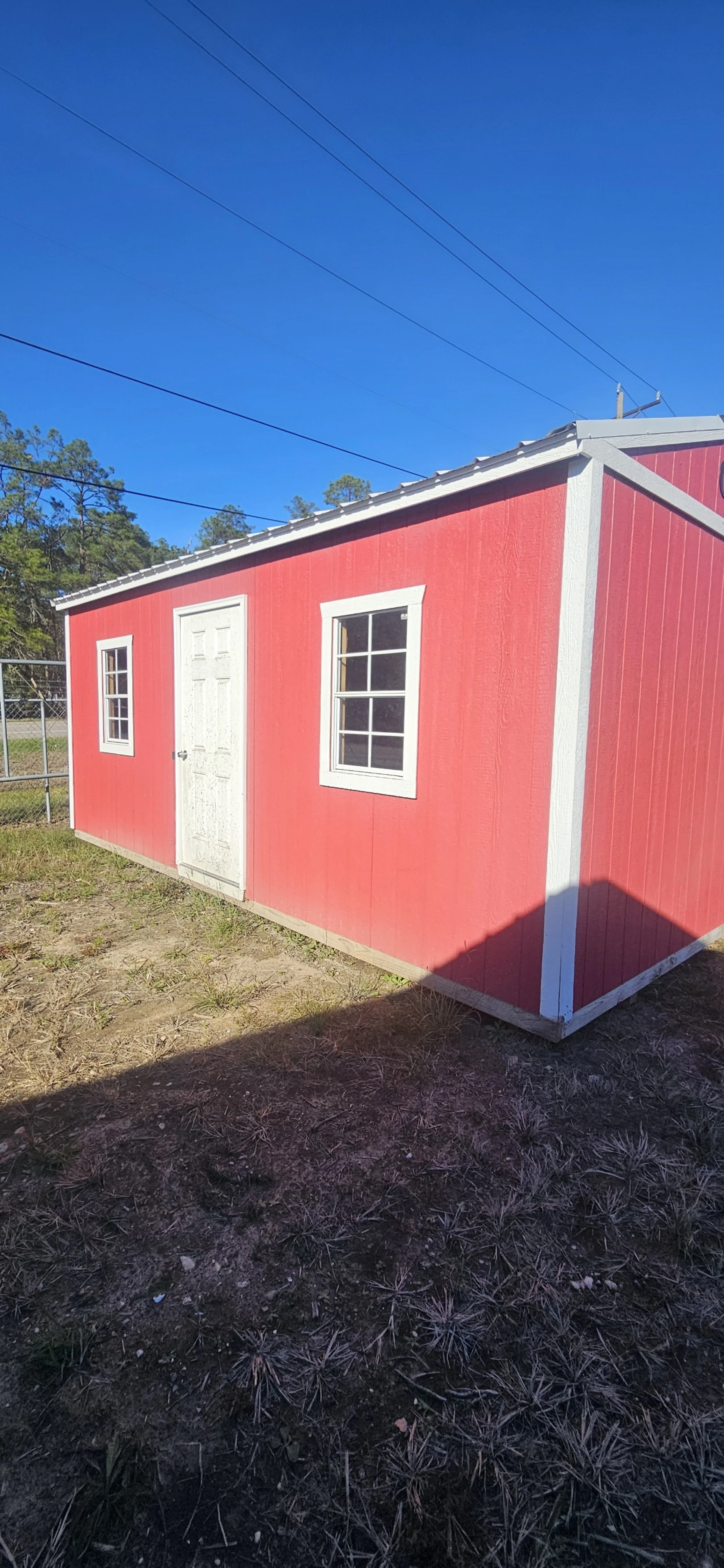 A small red shed with white trim, two windows, a door, set on a patch of dirt with a fence and trees in the background, under a clear blue sky.
