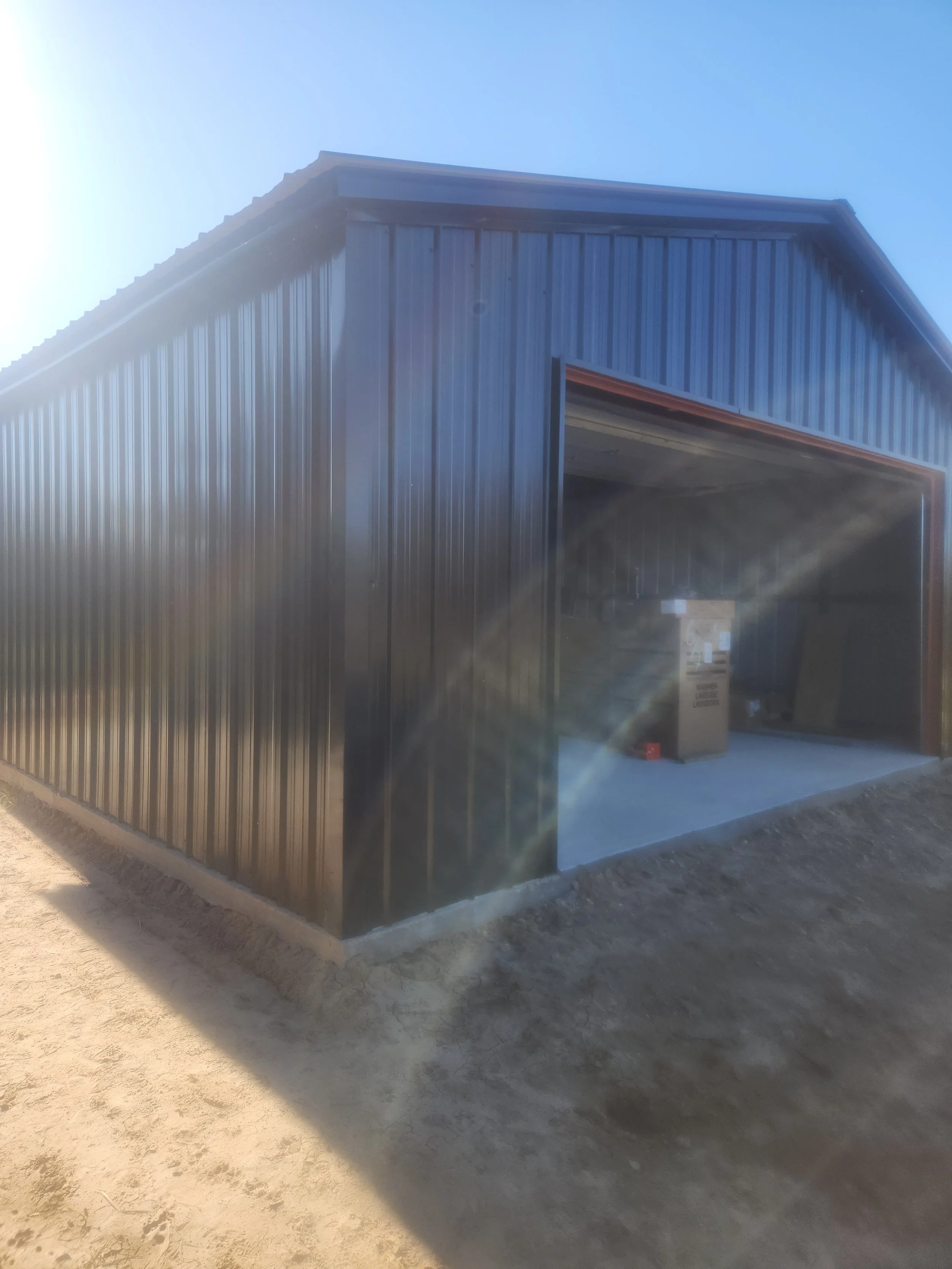 A black metal building with an open garage door, sunlight shining on it, and a concrete floor with a trash can inside.