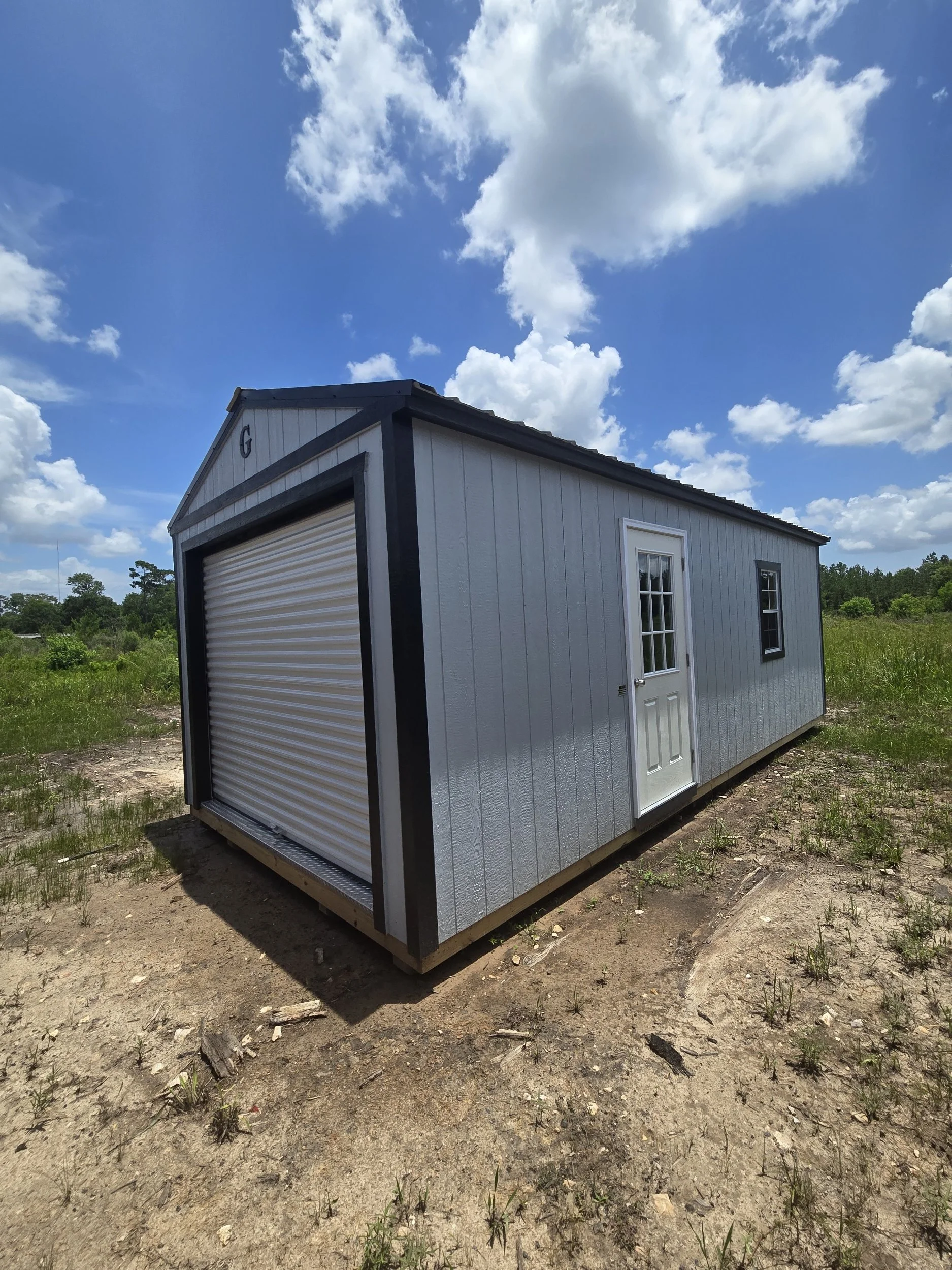 Small gray outdoor shed with a white door and a roll-up garage door, situated on a patch of dirt with grass, under a partly cloudy blue sky.