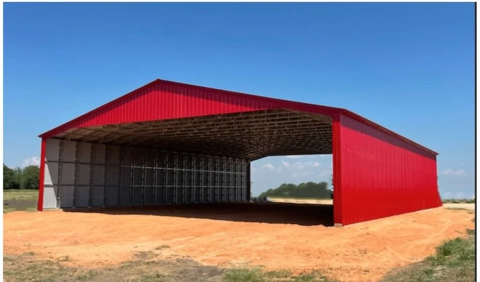 A large red metal storage shed with open front, situated on a dirt ground with some patches of grass, under a clear blue sky.