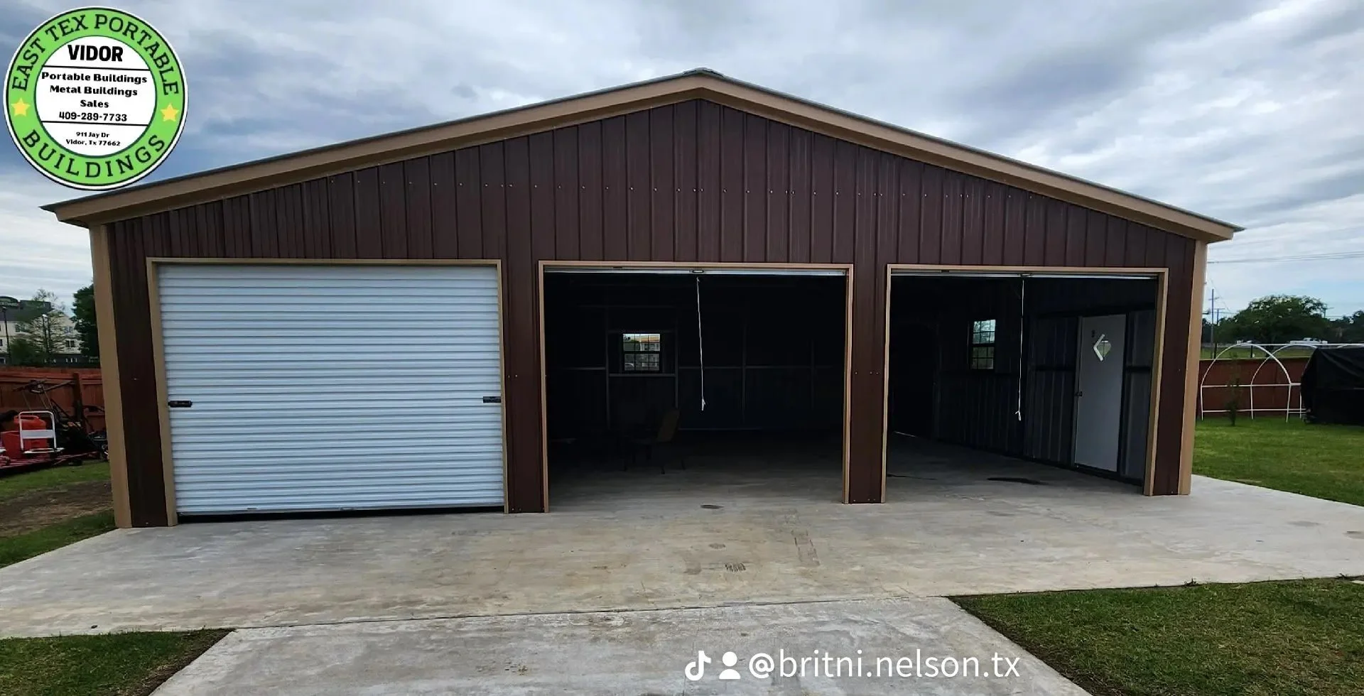 A new metal building with a brown exterior and a sloped roof, featuring two large garage doors—one closed, the other open—and an interior with visible framing and small window. The building is on a concrete slab with grass on either side, and a cloud