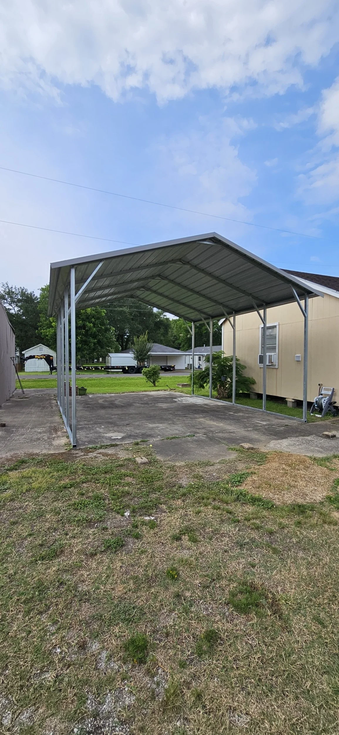 A carport with a metal roof and supporting beams, set on a concrete slab, in a residential yard with grass and a small tree, behind a house with beige siding. In the background are trees, other houses, and a partly cloudy sky.