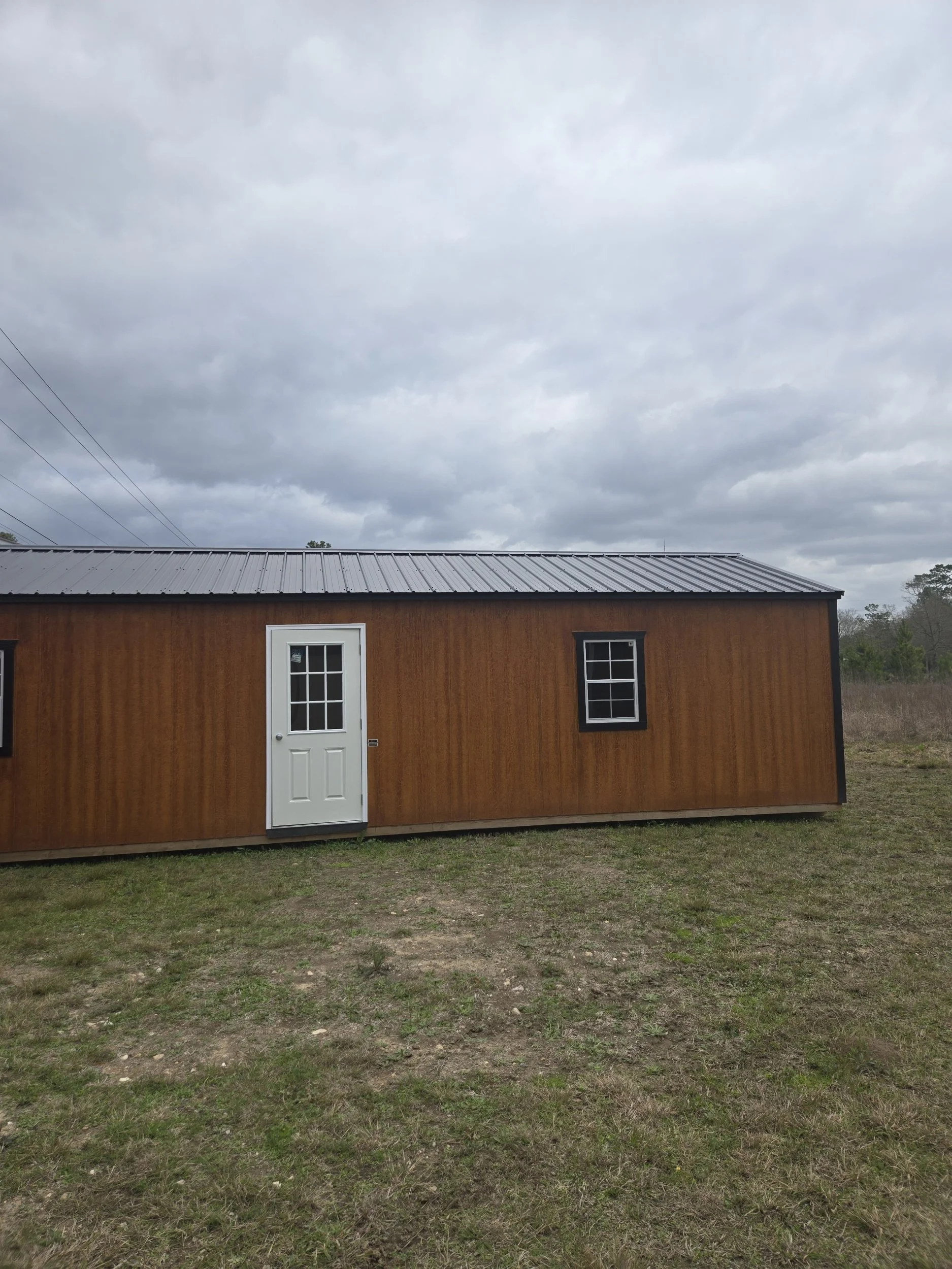 A small wooden building with a white door and two windows, located in an open grassy area under a cloudy sky.