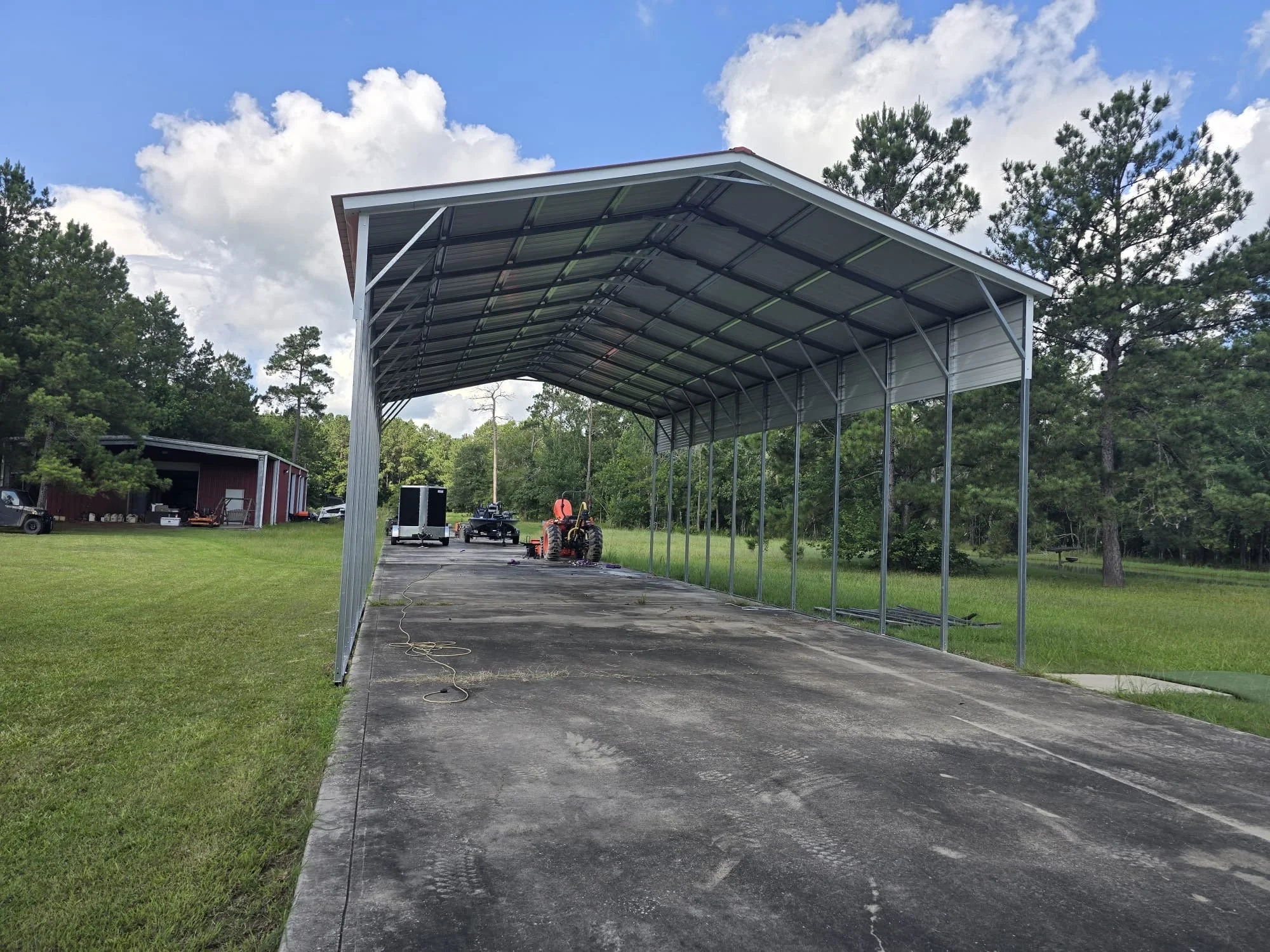 A metal carport structure being assembled on a concrete driveway in a rural setting with green grass and trees, with construction equipment and vehicles nearby on a partly cloudy day.