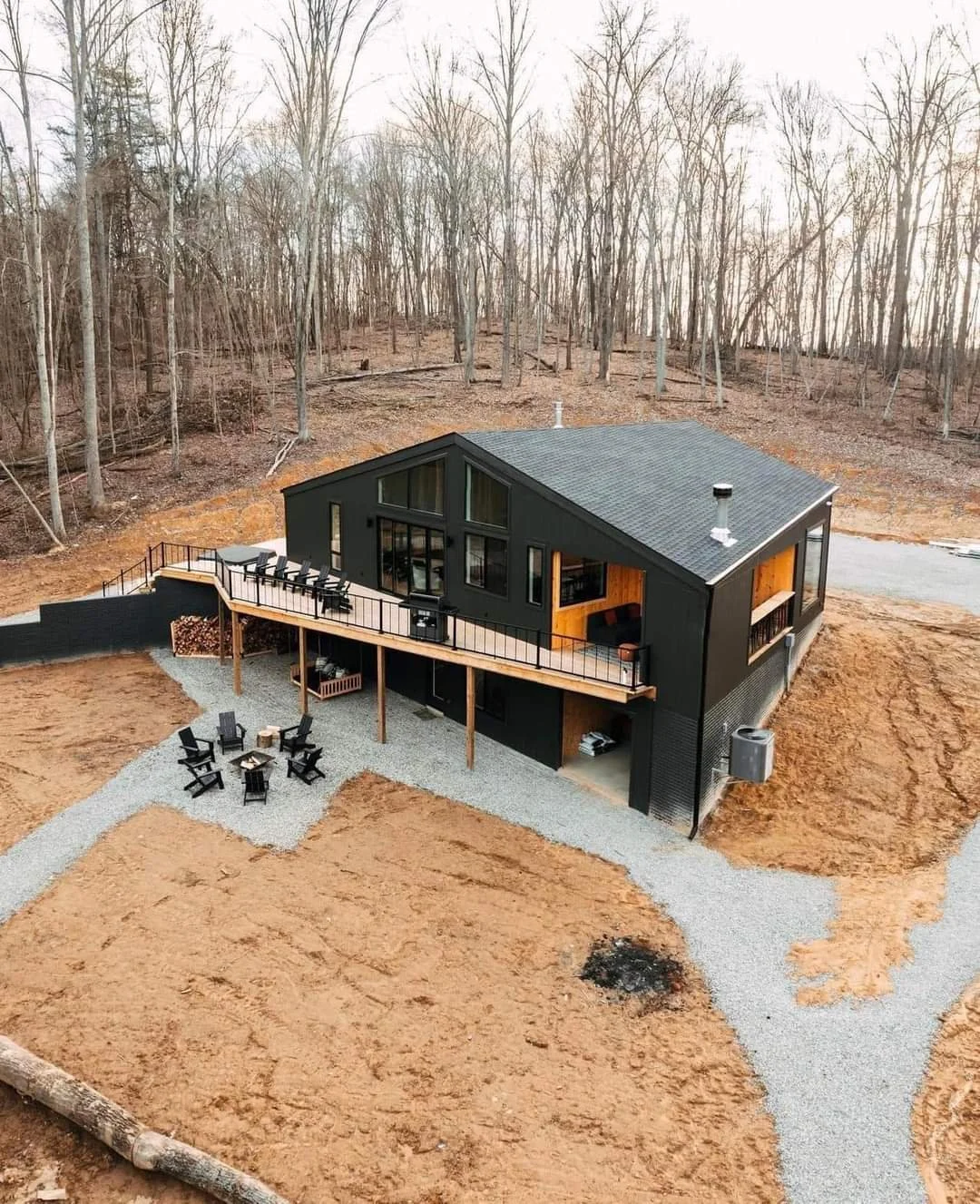 A modern black house with large windows and a sloped roof, set on a cleared wooded lot with a gravel driveway, outdoor seating area with Adirondack chairs, and a wooden balcony.