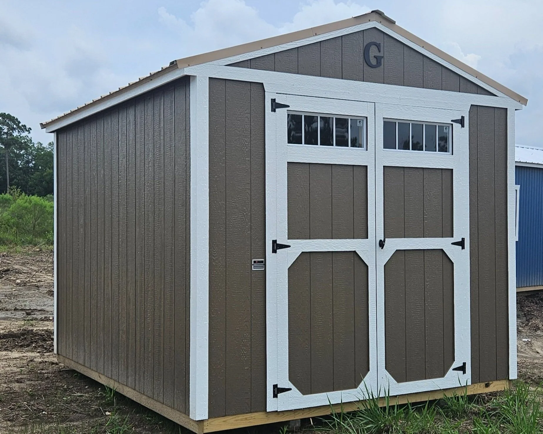 A small shed with brown panels and white trim, featuring double doors with black hinges, a small window at the top, and a gray roof, situated outdoors on grassy and dirt terrain under a cloudy sky.