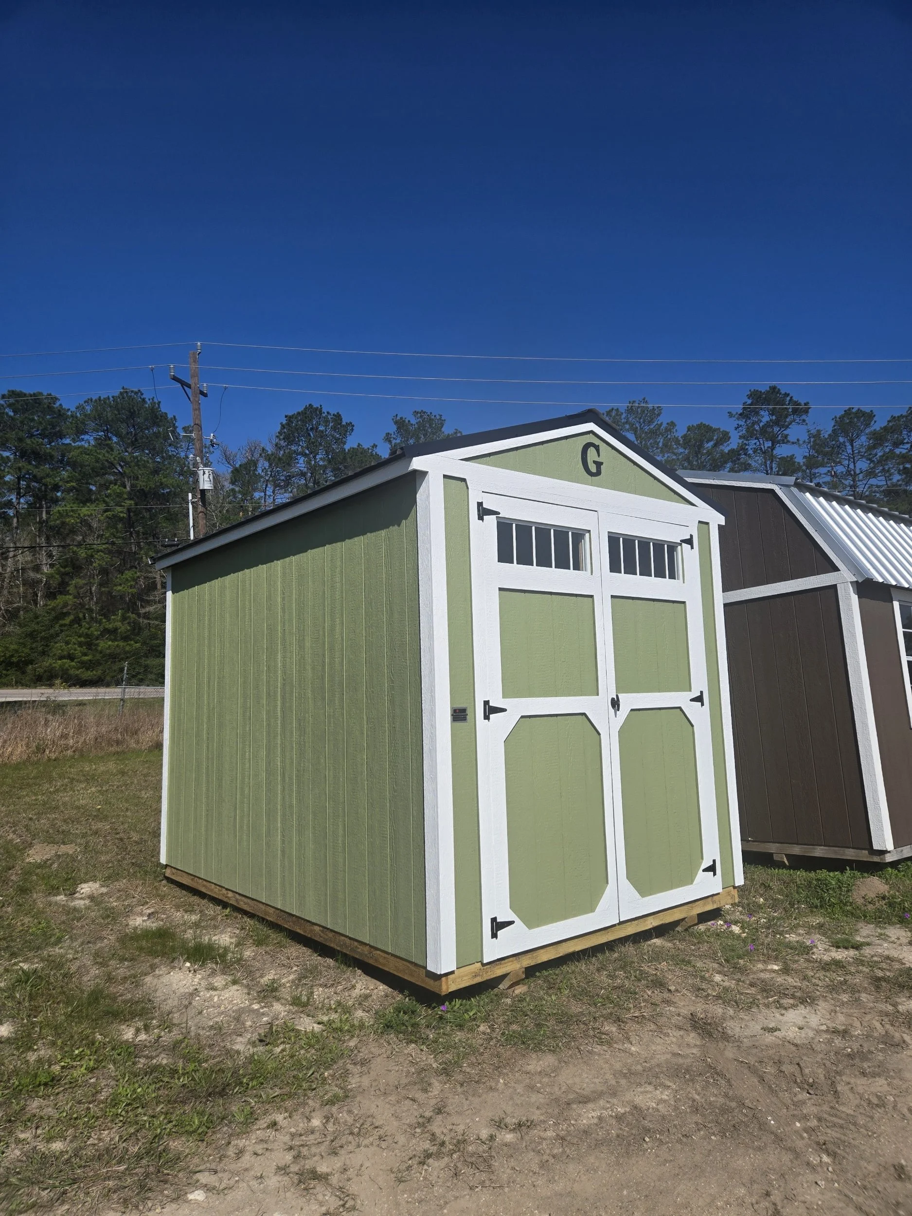 Green and white shed with a gable roof, located outdoors on a dirt patch, with trees and power lines in the background.