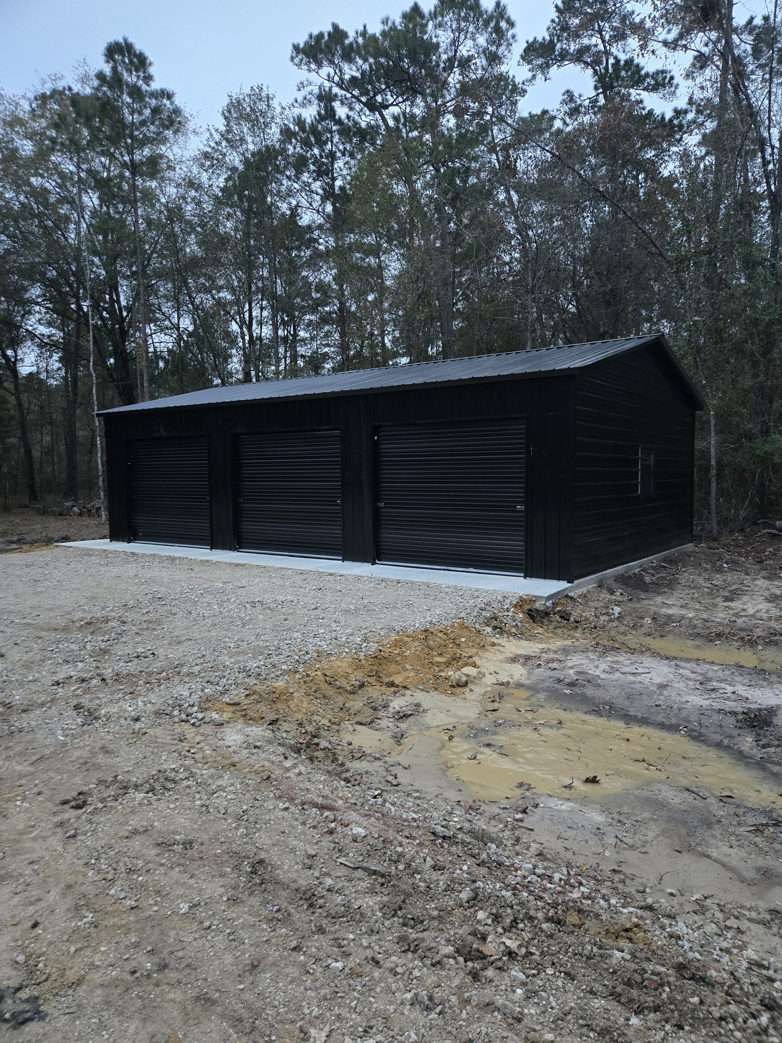 A black metal garage with three roll-up doors is situated on a concrete slab in a wooded area. The ground surrounding the garage is uneven with dirt, gravel, and small puddles.
