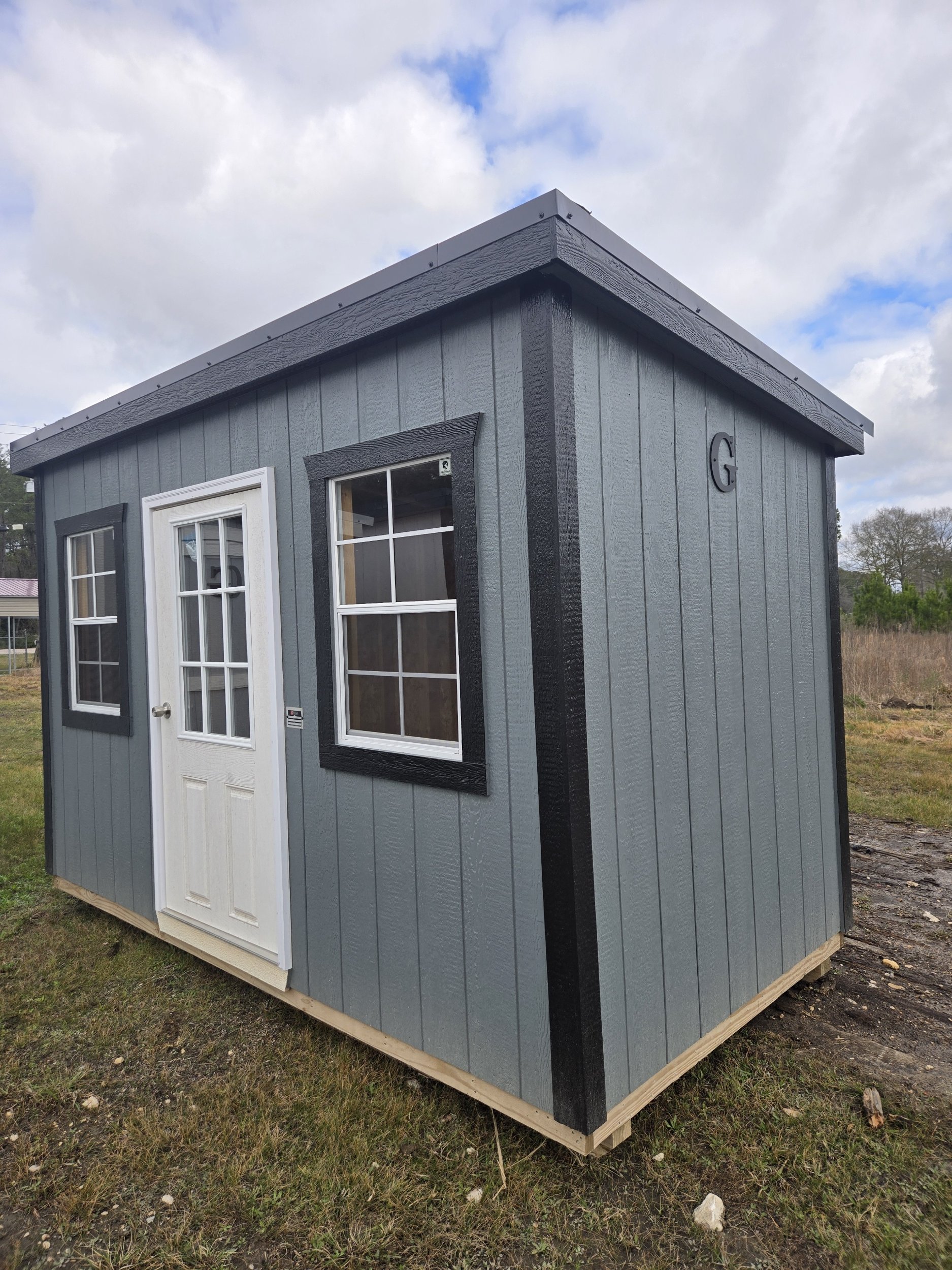 Small gray shed with white door and windows, black trim, and the letter G on the side, situated on a grassy area under a partly cloudy sky.