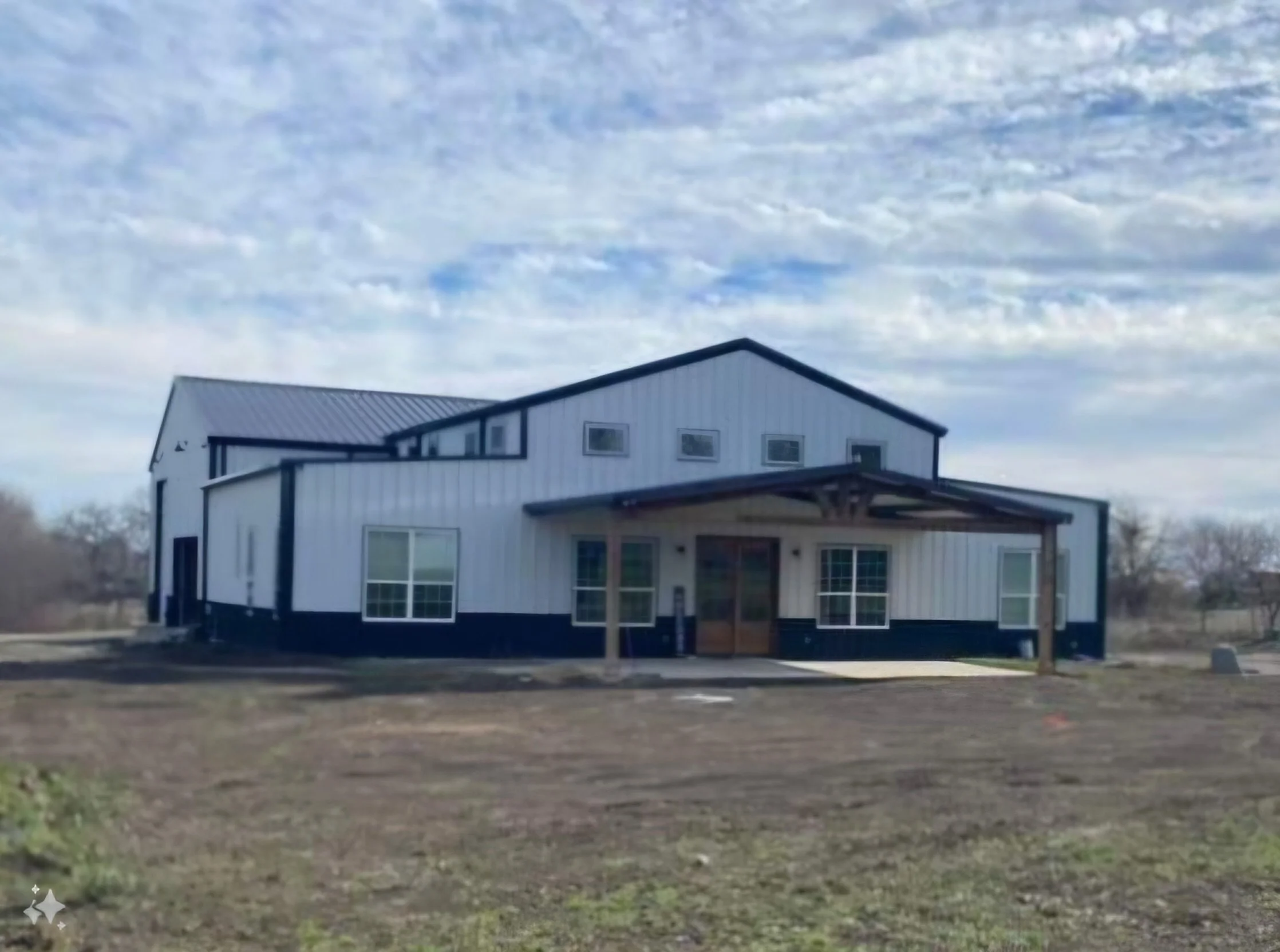 A two-story house with white siding and black trim, featuring a covered porch supported by wooden posts, surrounded by an open yard with sparse grass and trees in the background, under a partly cloudy sky.