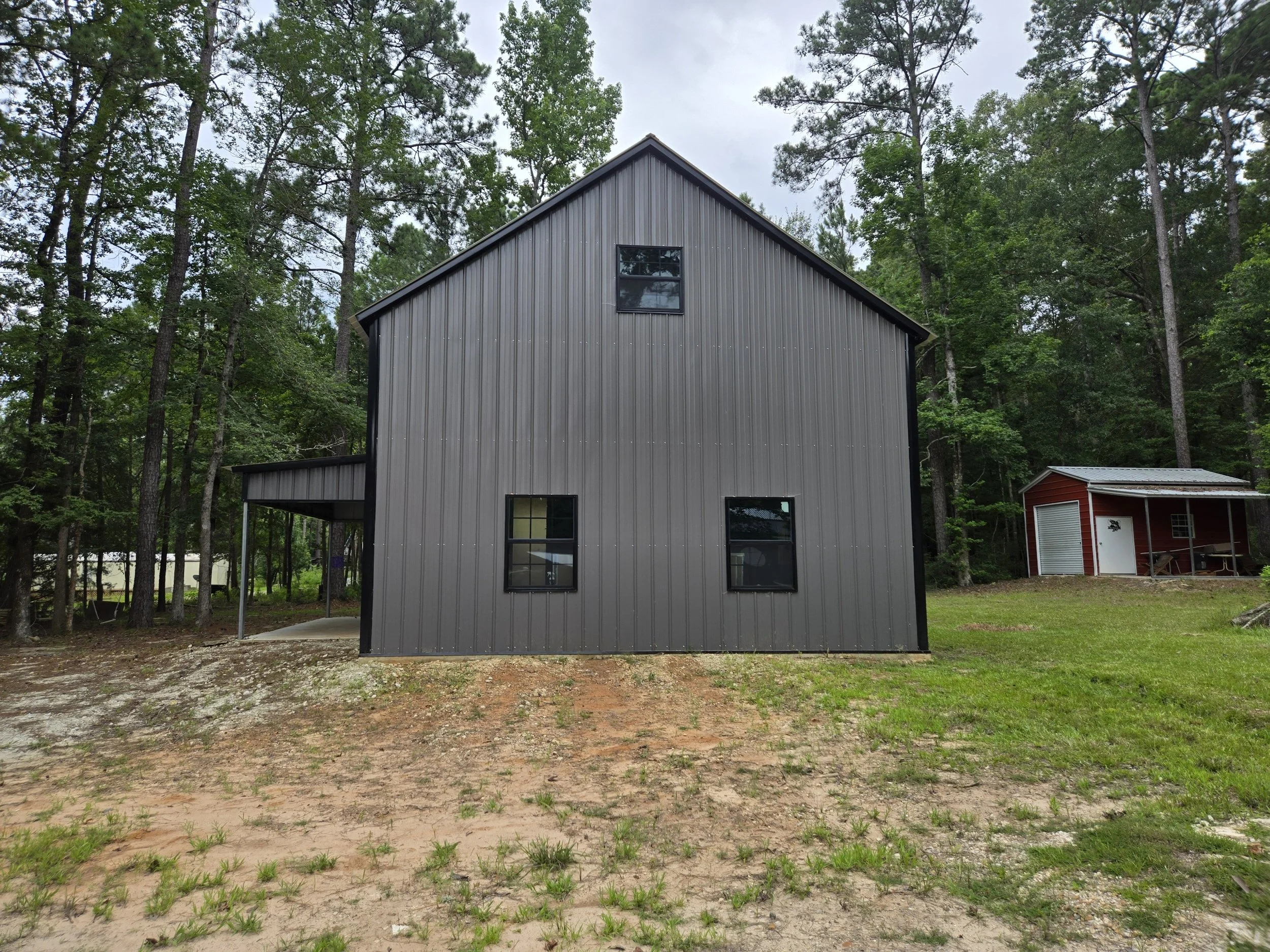 A modern gray metal house with black window frames on a slightly sloped terrain surrounded by trees, with a small red shed with a metal roof in the background.