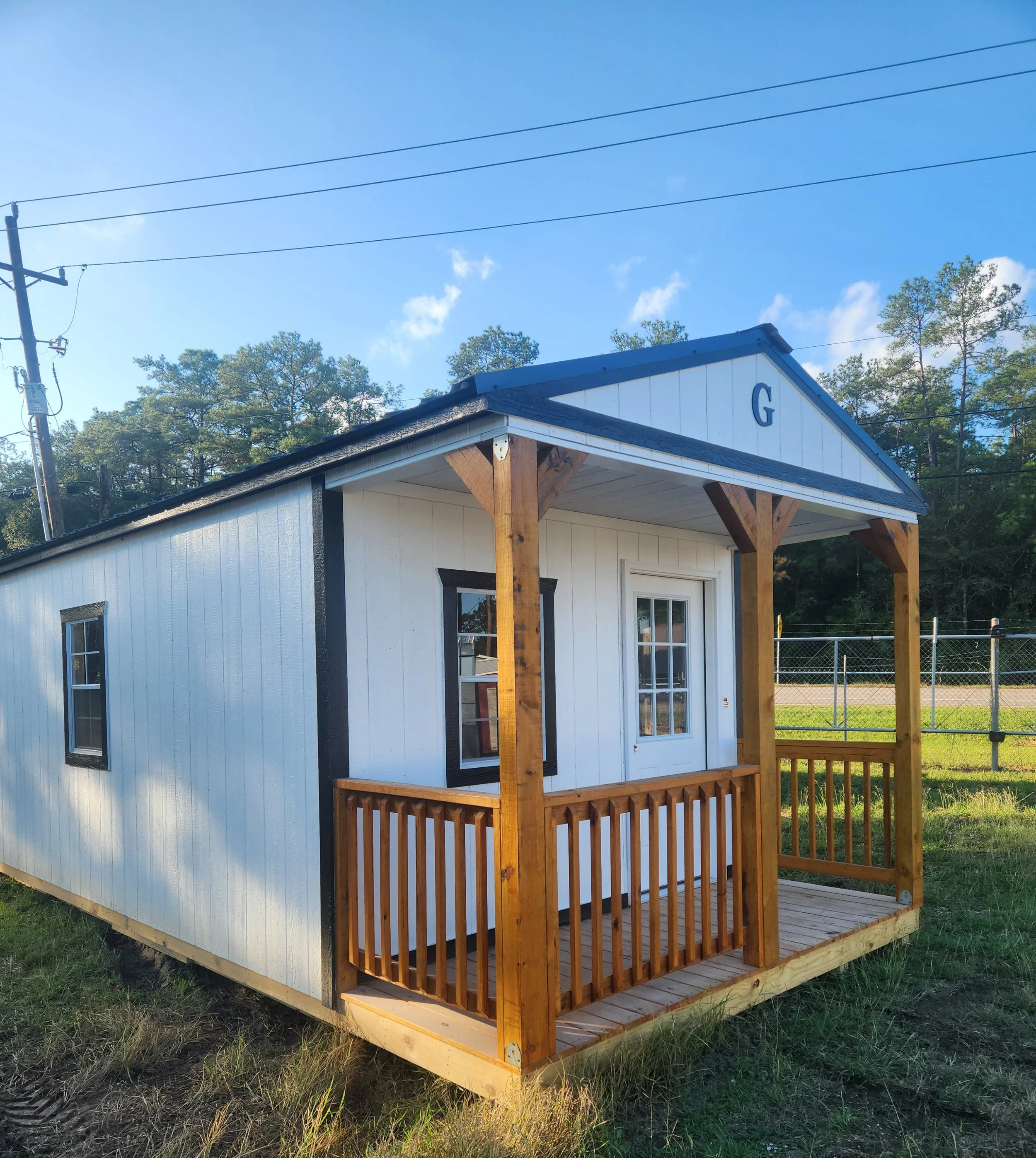 Small white and black tiny house with a front porch, wooden railing, and a peaked roof with a letter G on it, set in a grassy area with trees and a chain-link fence in the background.