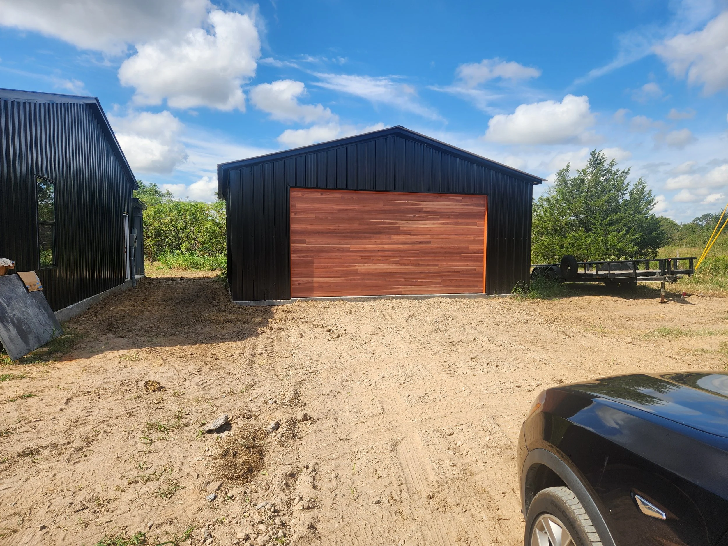 Black metal building with a large wooden garage door, surrounded by dirt and some green trees in the background, under a partly cloudy sky.