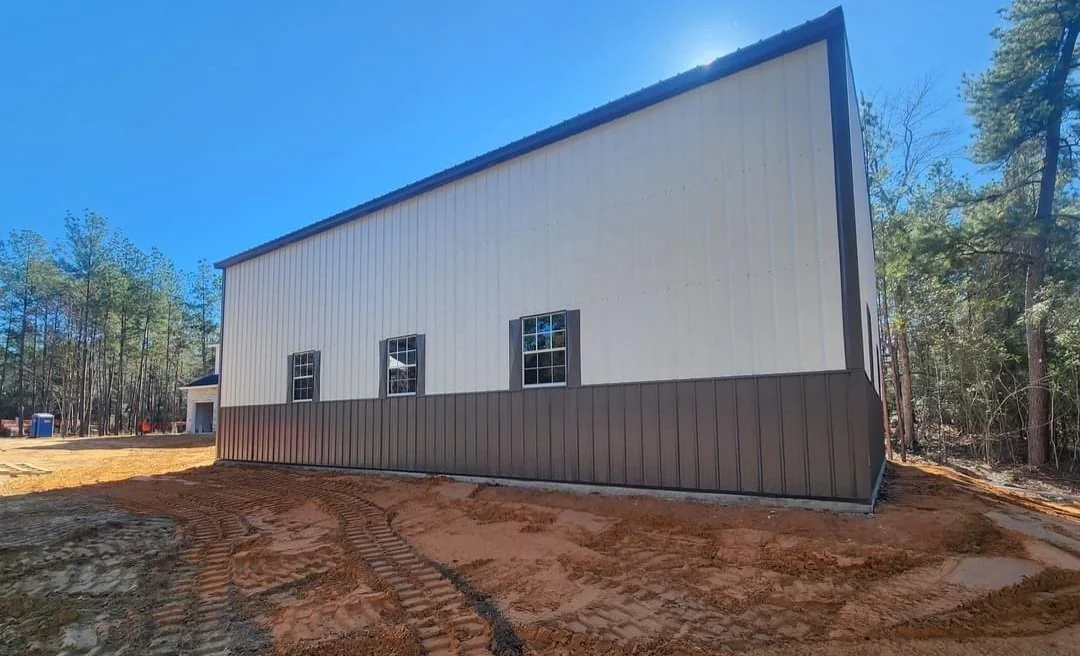 Newly constructed metal building with white and brown siding, three windows, surrounded by dirt with tire tracks, and trees in the background.