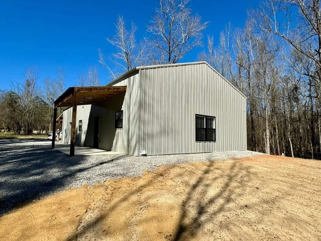 A metal building with a sloped roof, black windows, and a small covered porch, situated on a gravel driveway and surrounded by leafless trees with a clear blue sky overhead.