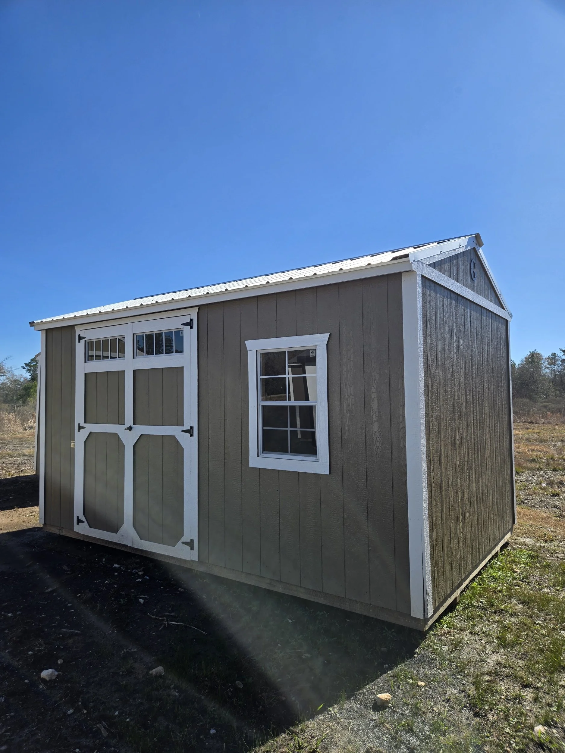 A small tan shed with white trim and double doors, a window, situated outdoors on a clear day with a blue sky.