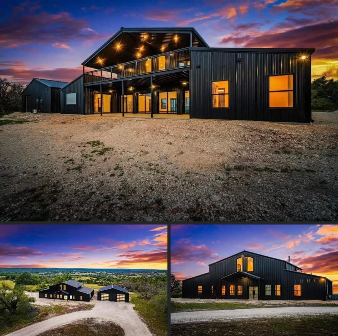 Modern black metal house with large windows and a deck, set in a rural area at sunset with a colorful sky.