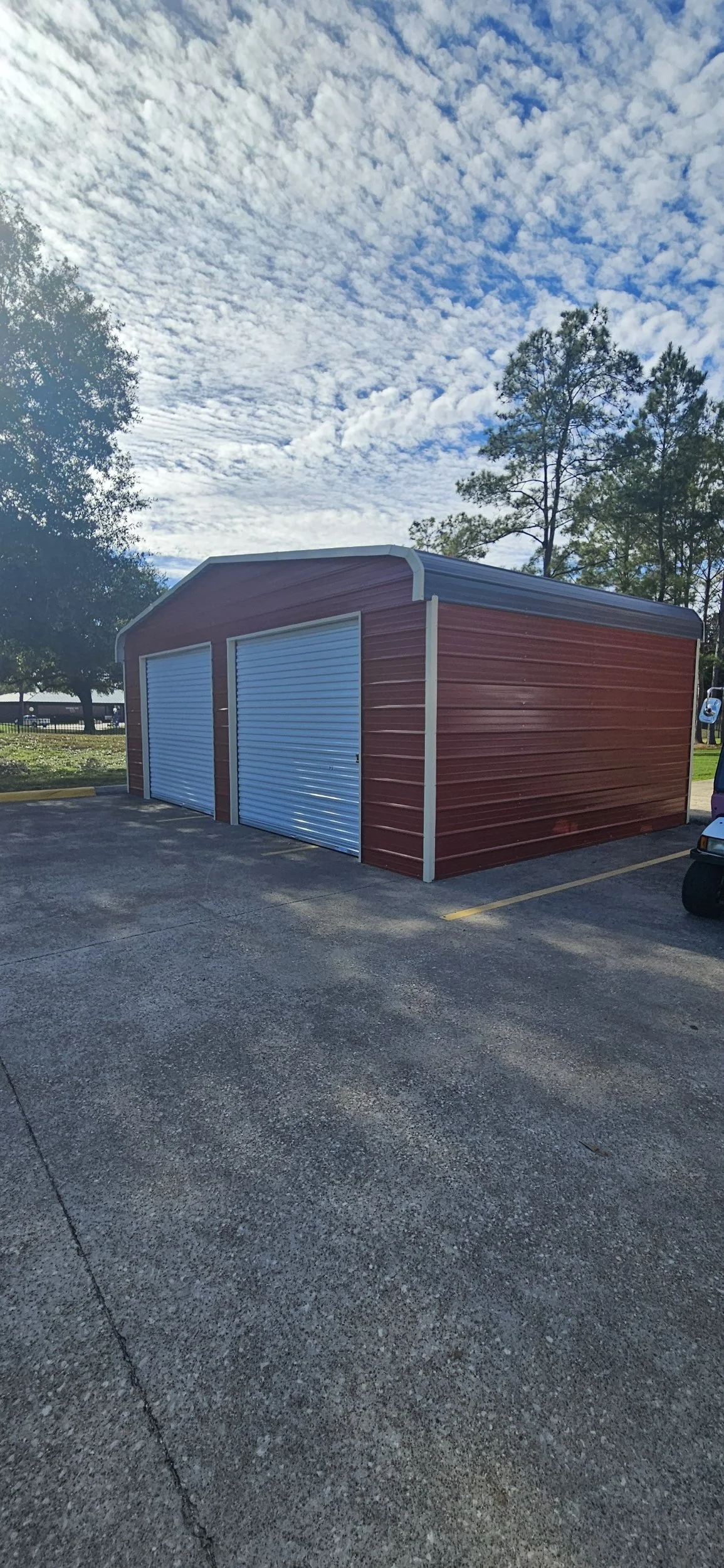A red metal storage building with two roll-up doors, situated in a parking lot under a partly cloudy sky, with trees in the background.