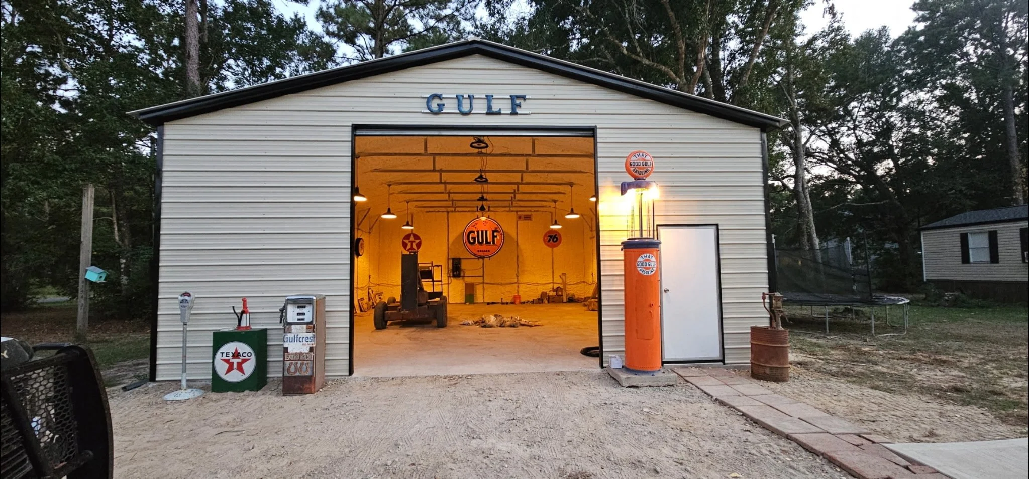 A garage with an open door labeled 'GULF'. Inside, vintage Gulf gas station signs and an old gas pump are visible. Outside, there are vintage gas station accessories, a tire changer, and a trampoline nearby.