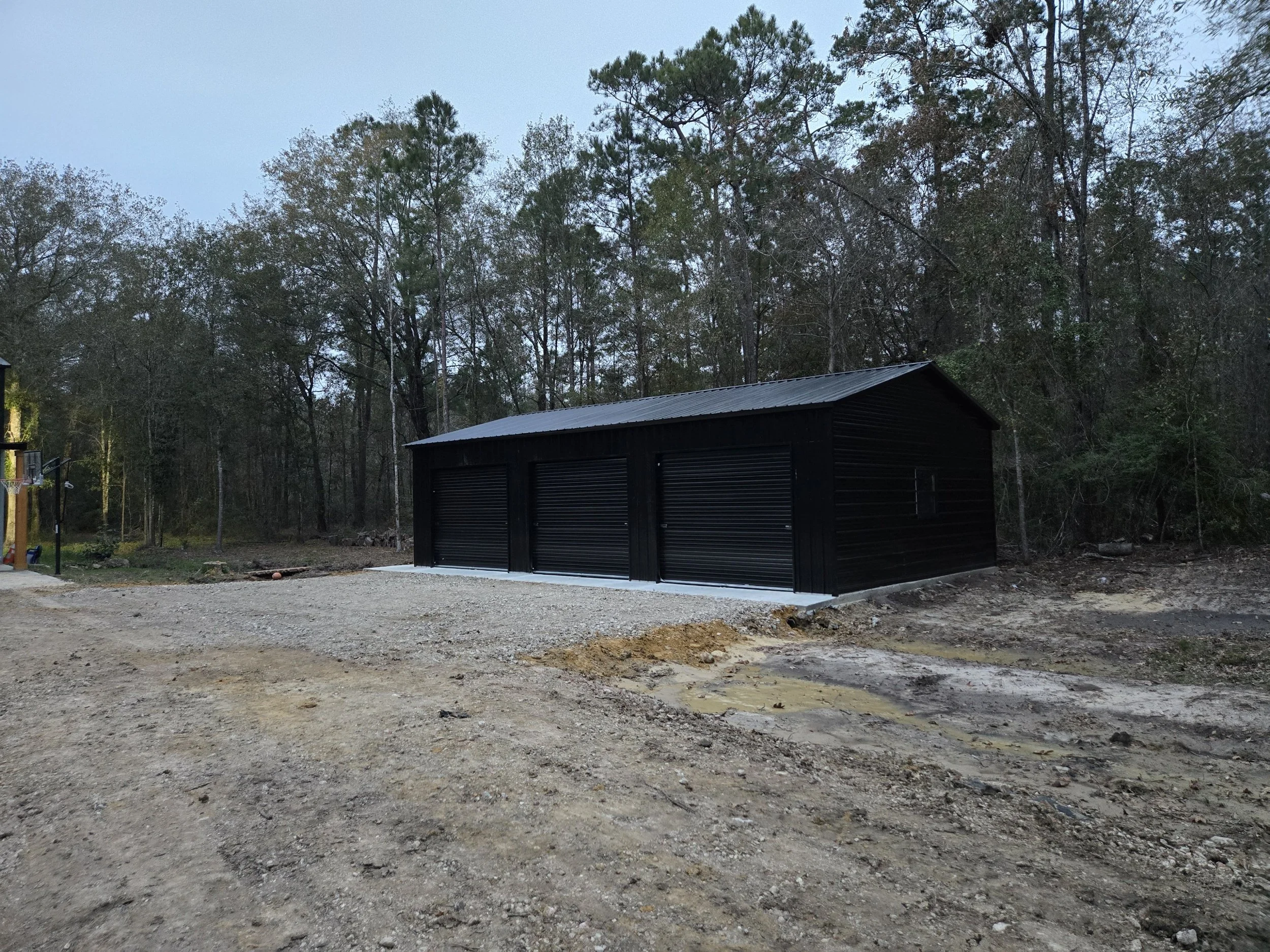A black metal garage with three roll-up doors located in a gravel area, surrounded by trees and a wooded background, during twilight.