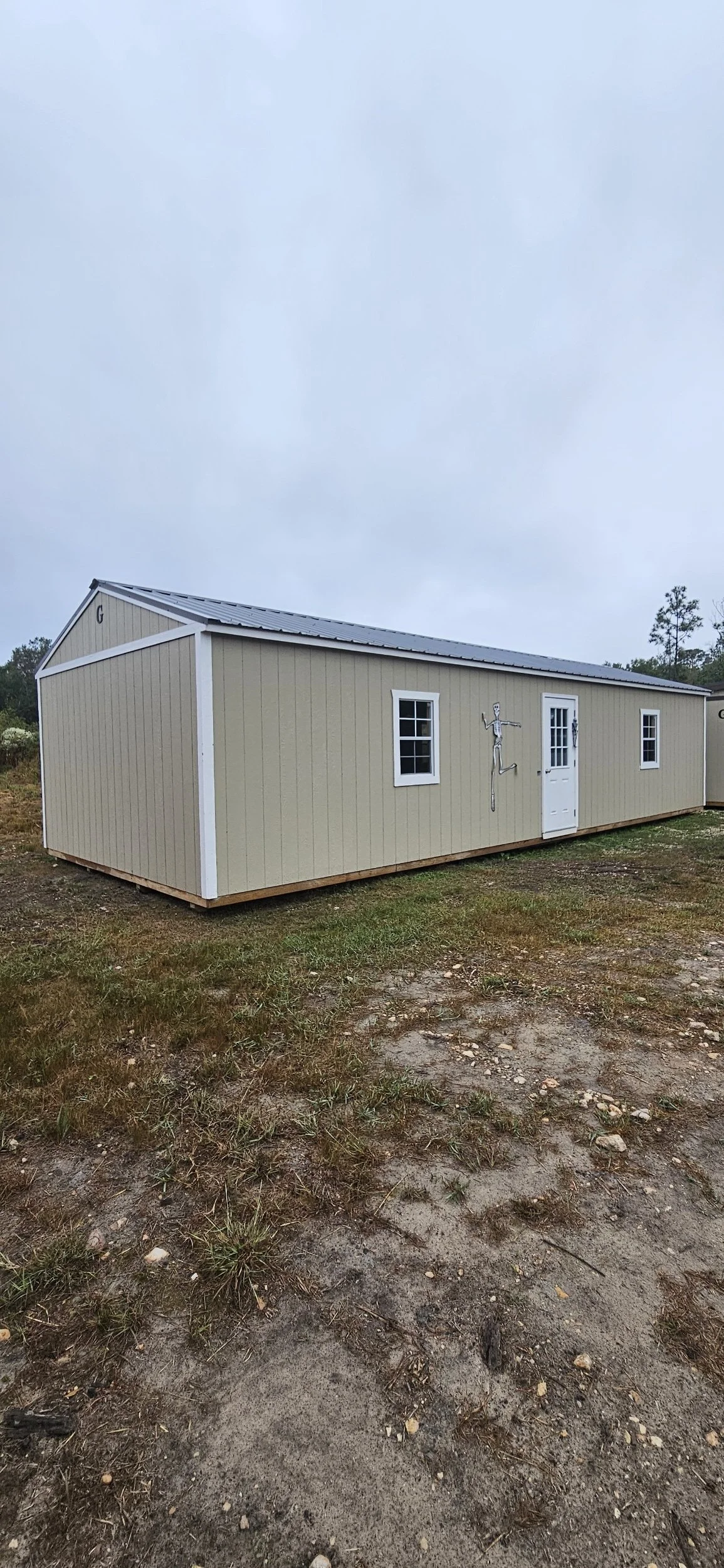 A beige mobile home with white trim, two windows, a door, and a skeleton decal on the side, situated on a patch of dirt and grass under a cloudy sky.