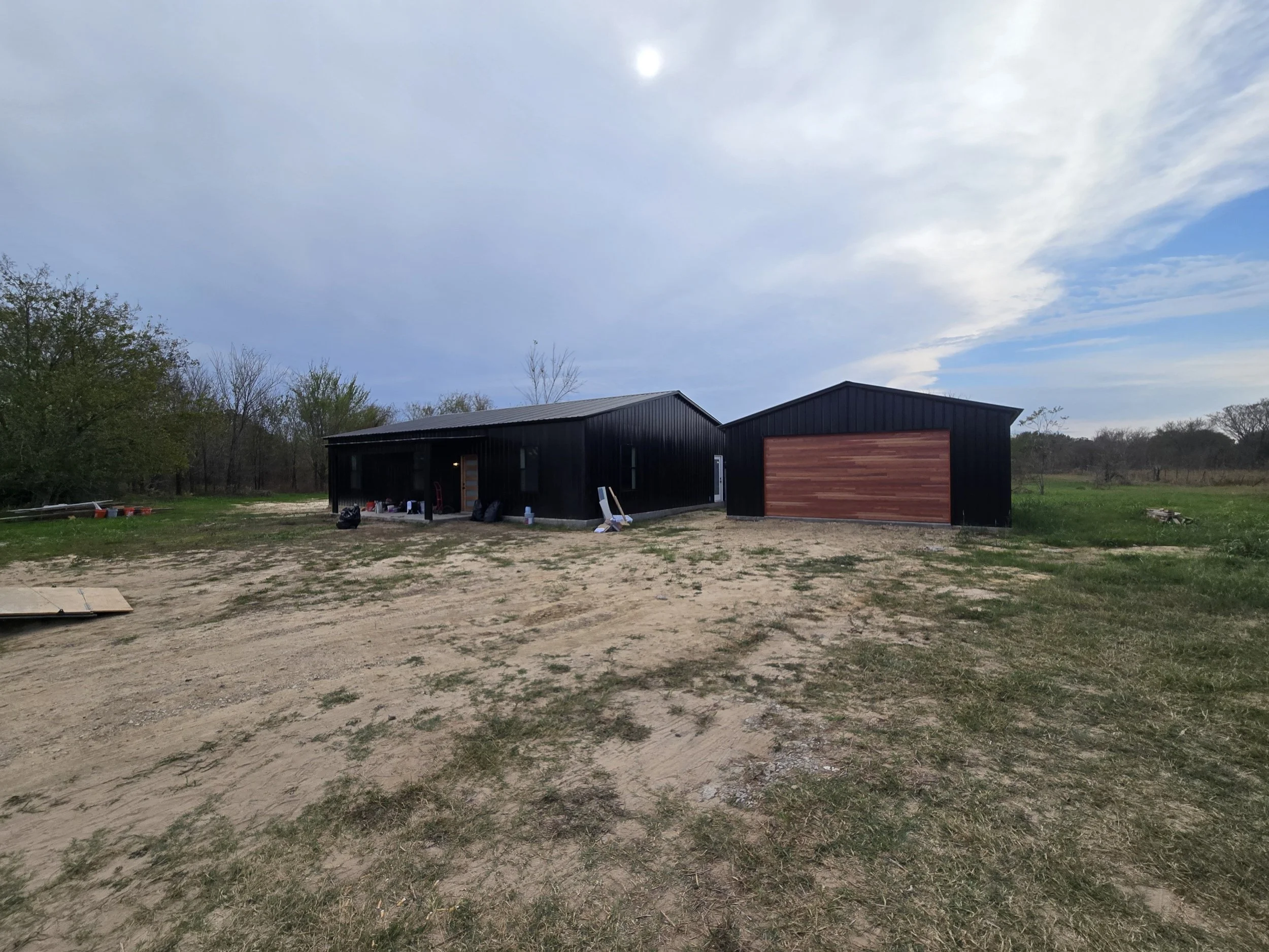 A black metal building with a wooden garage door, set in a rural area with sparse grass and dirt, against a backdrop of trees and a partly cloudy sky.