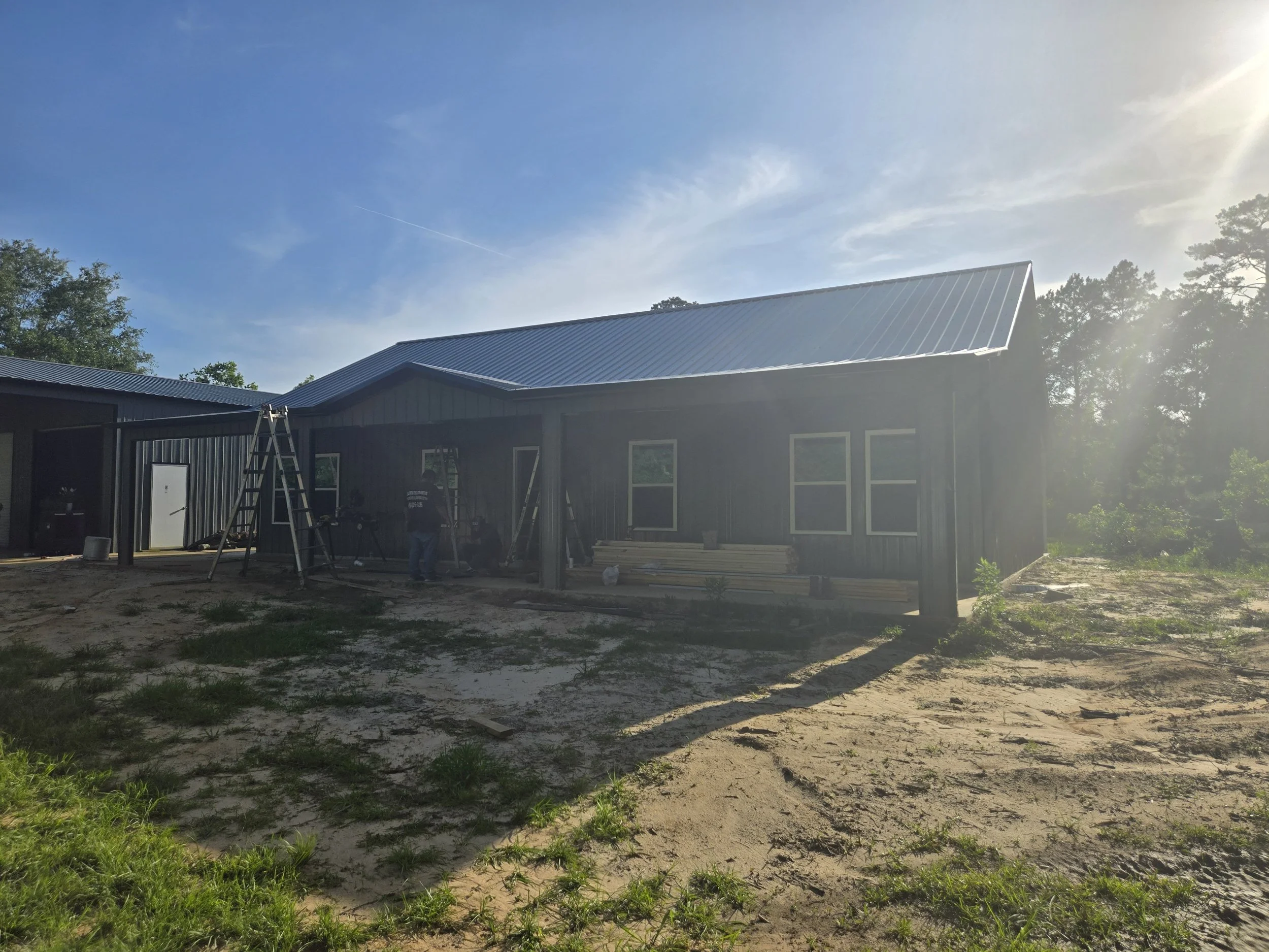 Under construction house with metal roof, fenced yard, and construction workers working on the front porch.