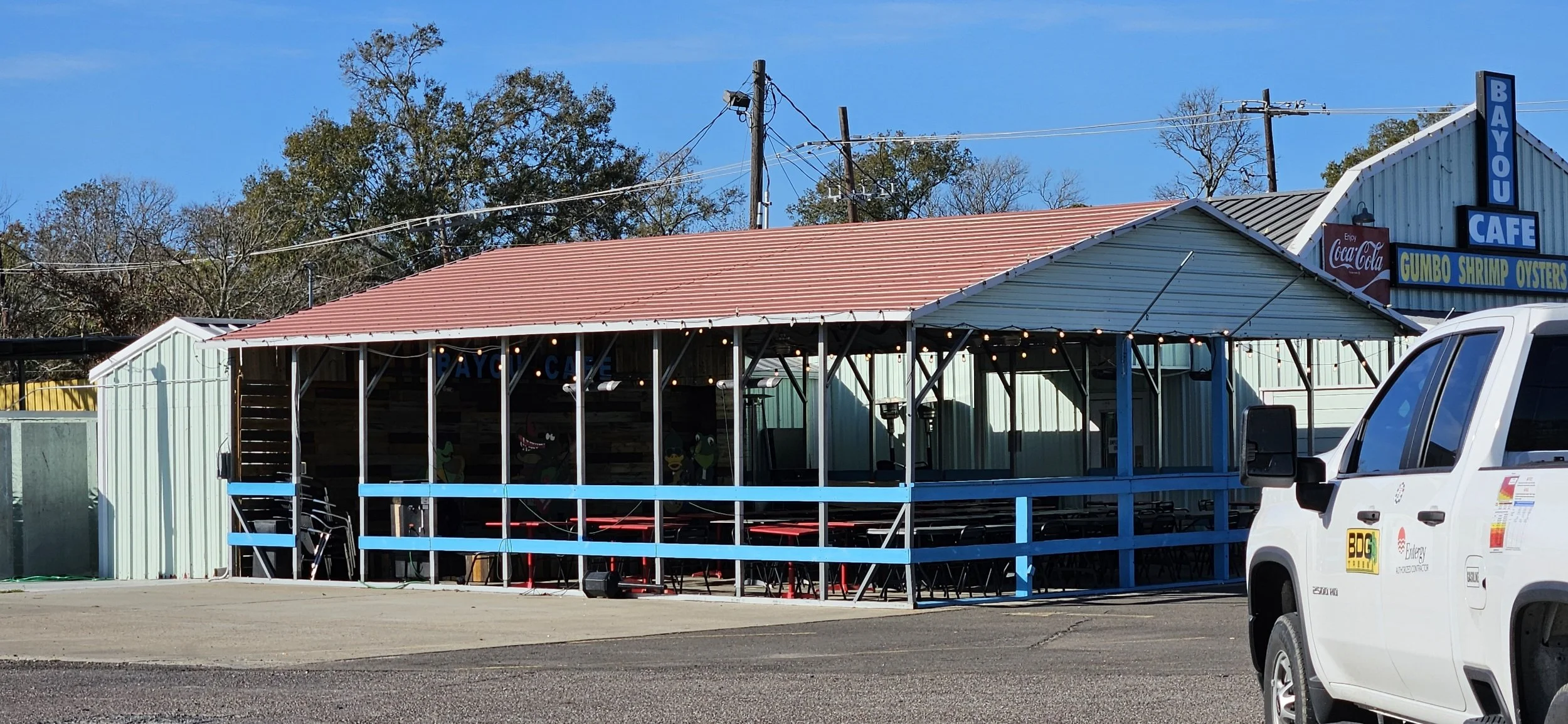 Exterior of a seafood restaurant with a screened outdoor seating area, blue railing, and a sign indicating 'Bayou Cafe' and 'Gumbo Shrimp Oysters', with a white vehicle parked in front and trees in the background.