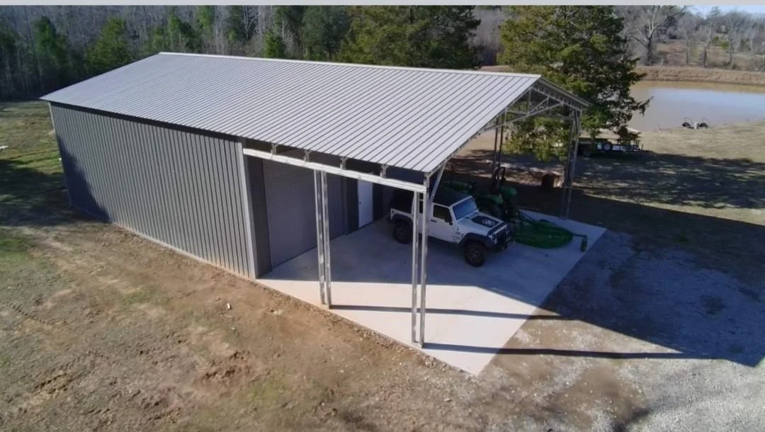 A metal storage shed with a sloped roof, open front, parked white truck, and a green tractor inside on a concrete pad, overlooking a pond surrounded by trees.