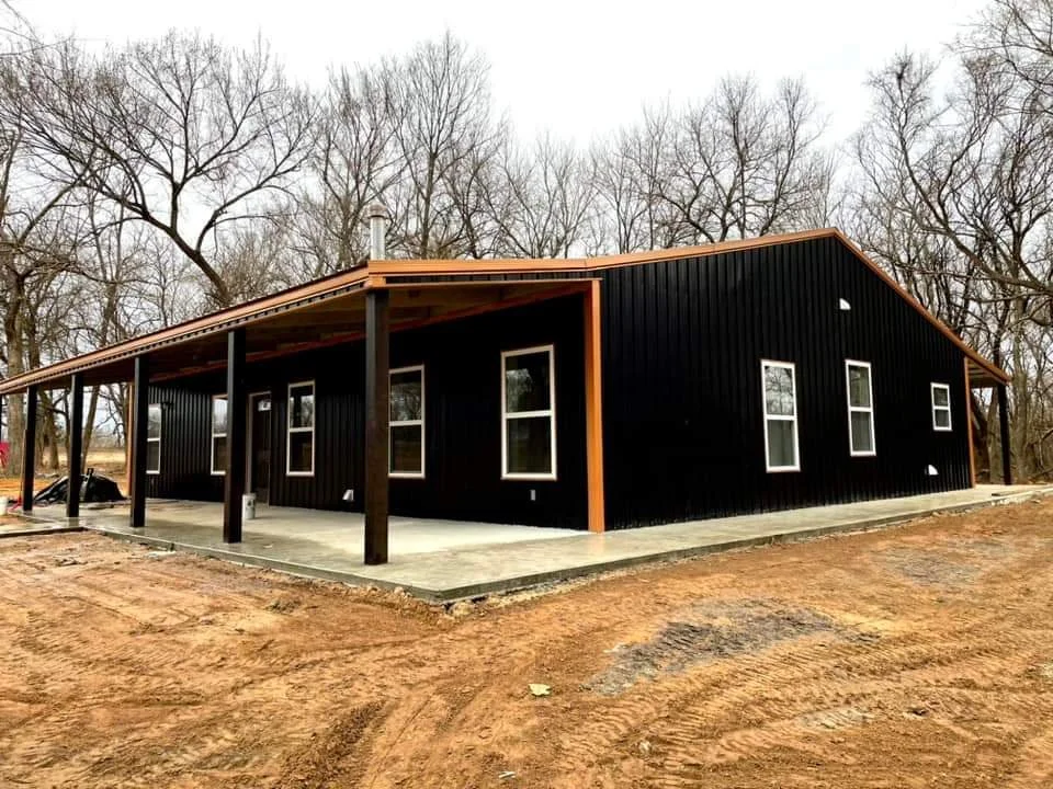 Newly constructed house with black metal siding, wooden trim, multiple white-framed windows, and a covered porch, situated on a dirt lot with leafless trees in the background.