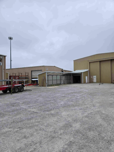 Industrial warehouse buildings with a gravel lot, a trailer on the left, and cloudy sky overhead.