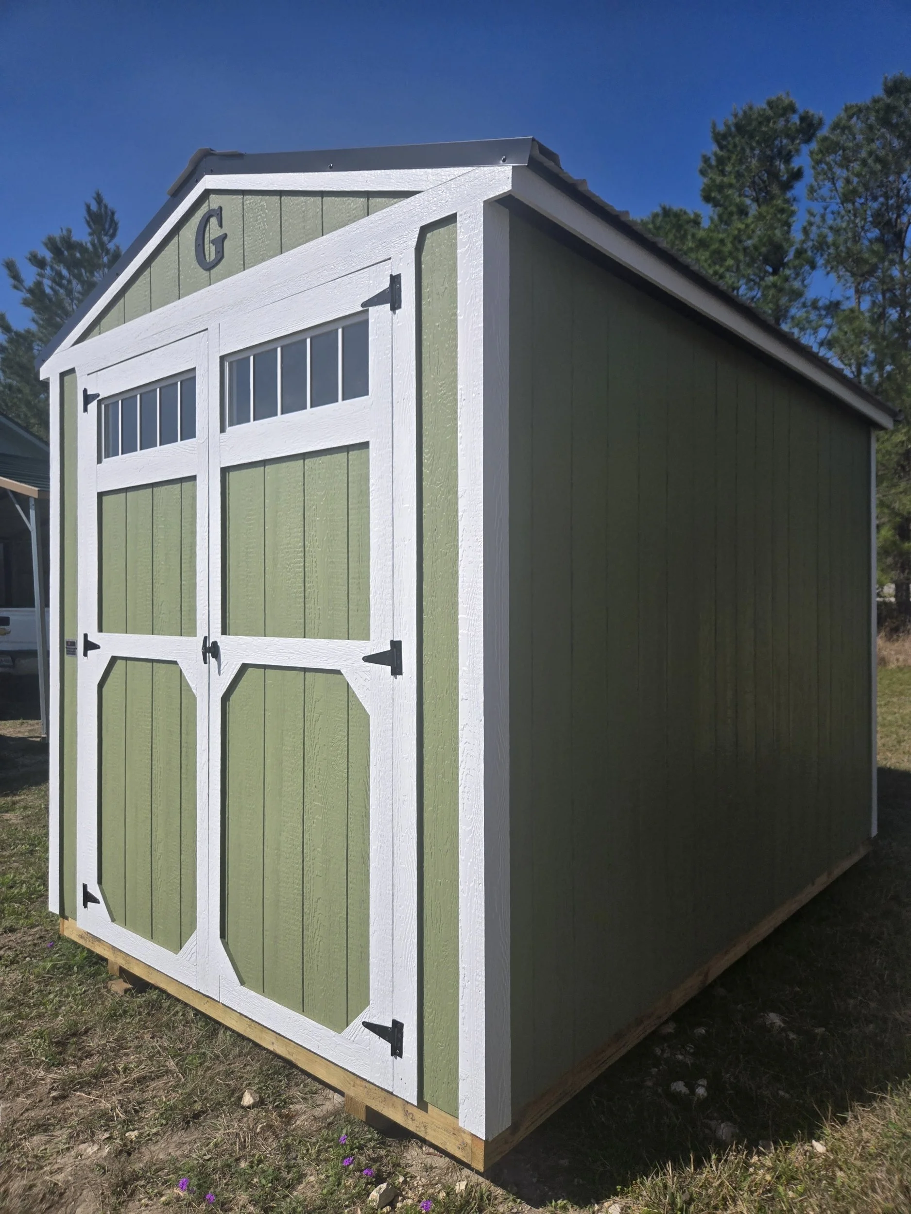 Green shed with white trim and black hardware, set outdoors on a wooden platform with trees and blue sky in the background.