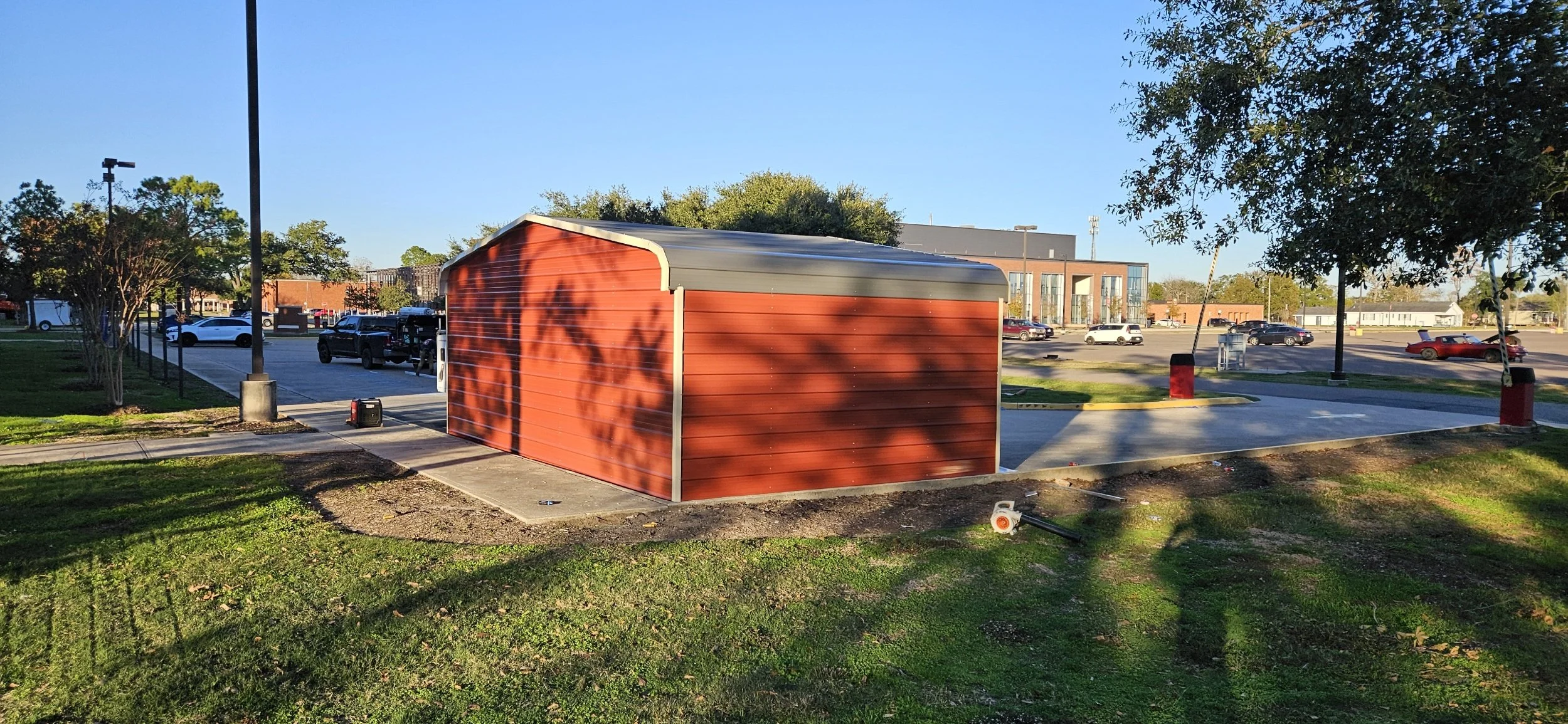 A red outdoor storage shed with a gray curved roof in a parking lot, surrounded by trees and parked cars, with a mostly clear blue sky in the background.