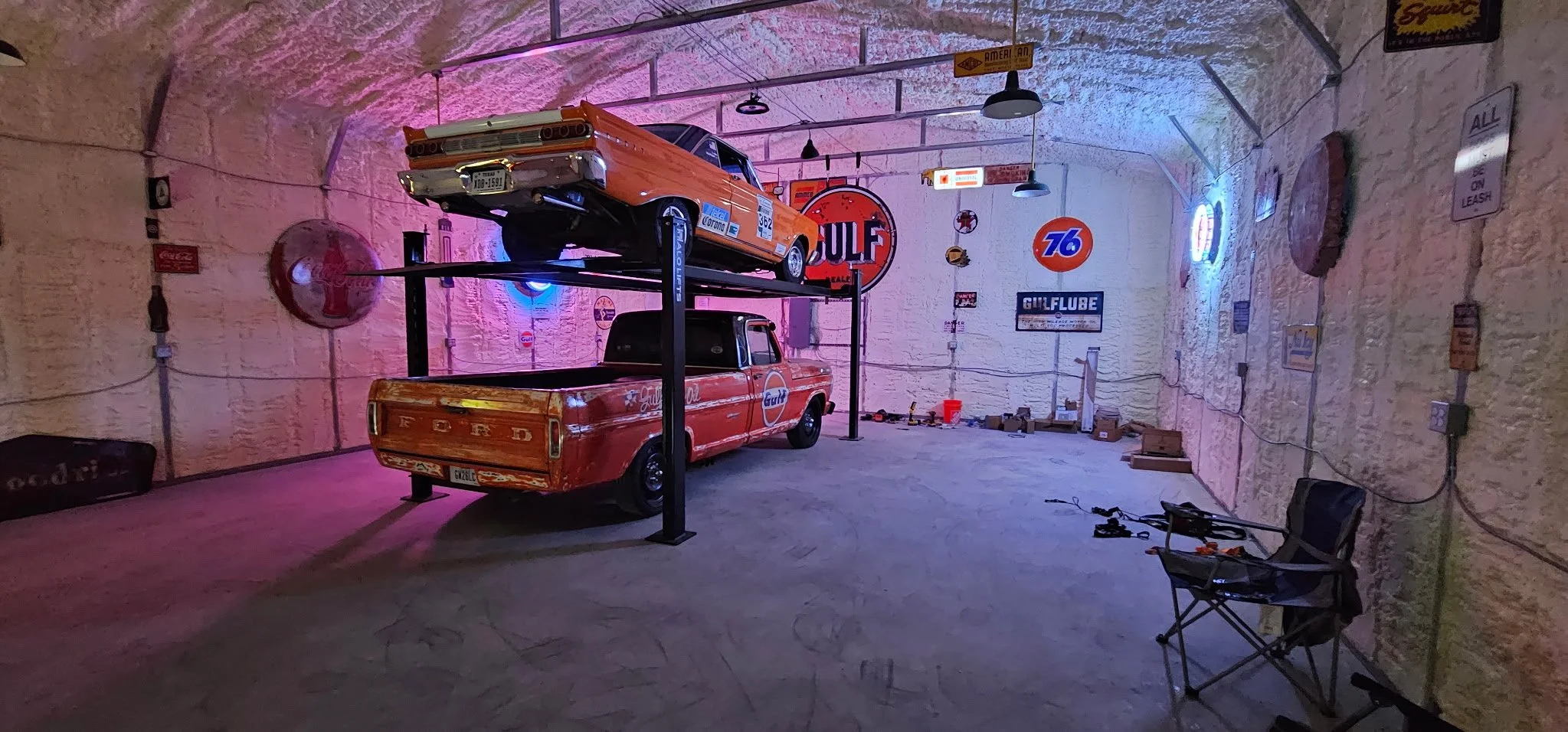 A room with vintage car memorabilia including two vintage cars, various neon signs, and vintage advertisements on the walls. The room has stone walls and is decorated with a neon pink and blue glow.