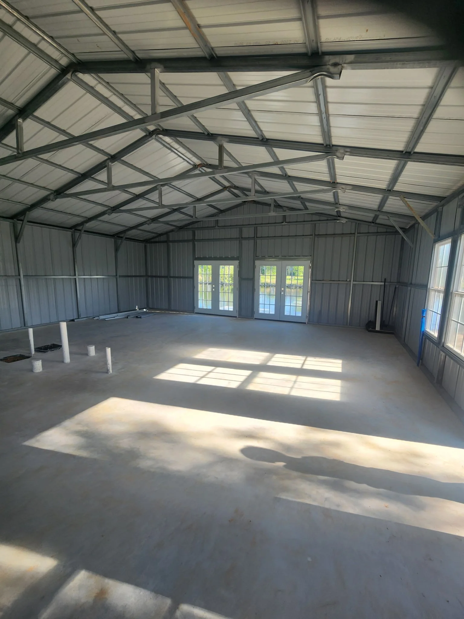 Empty metal garage or workshop with concrete floor, metal walls, and ceiling, with sunlight coming through glass double doors and windows, some pipes and construction materials on the floor.