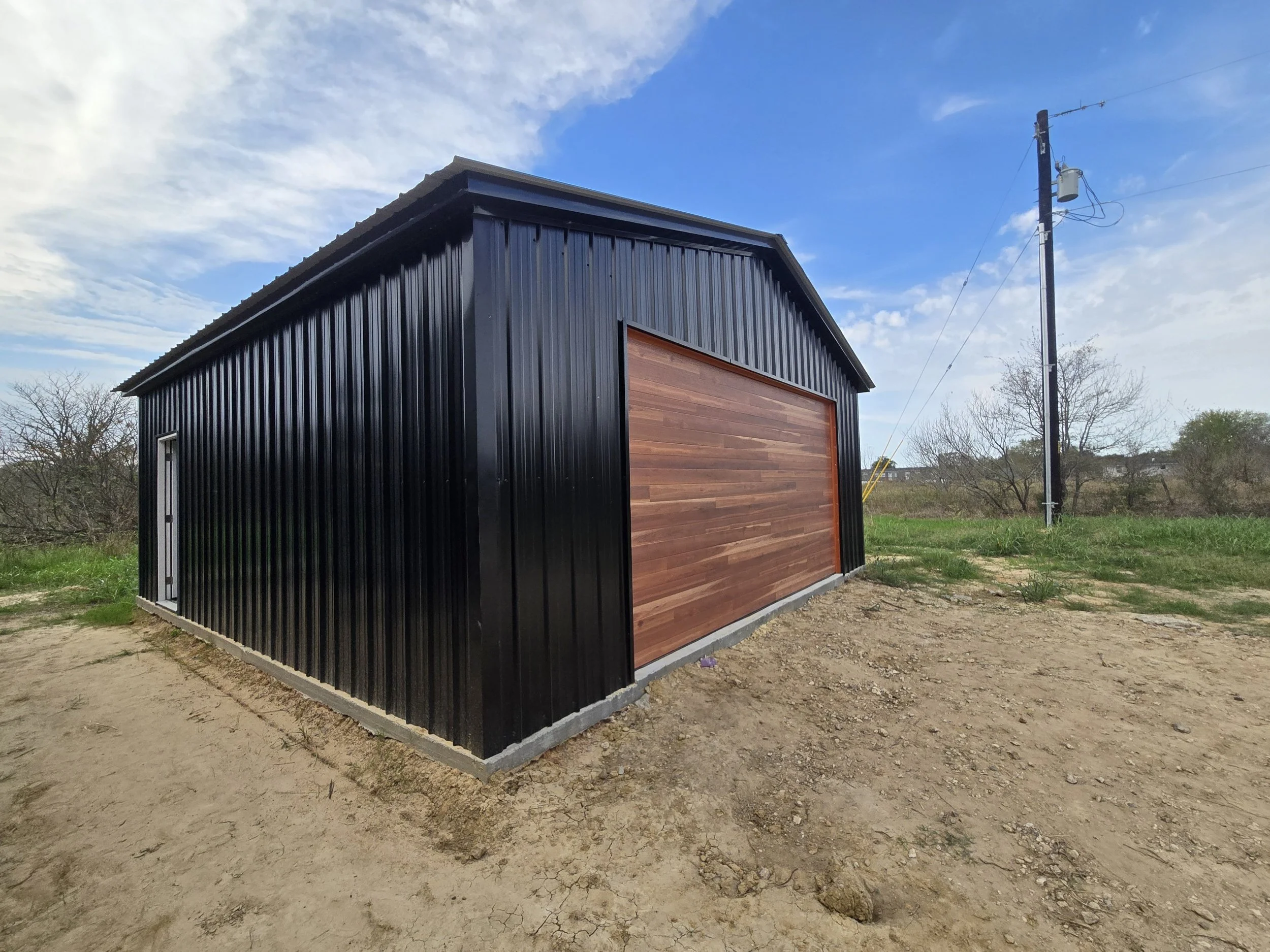 Black metal shed with a wooden garage door in an open grassy area under a blue sky with scattered clouds.