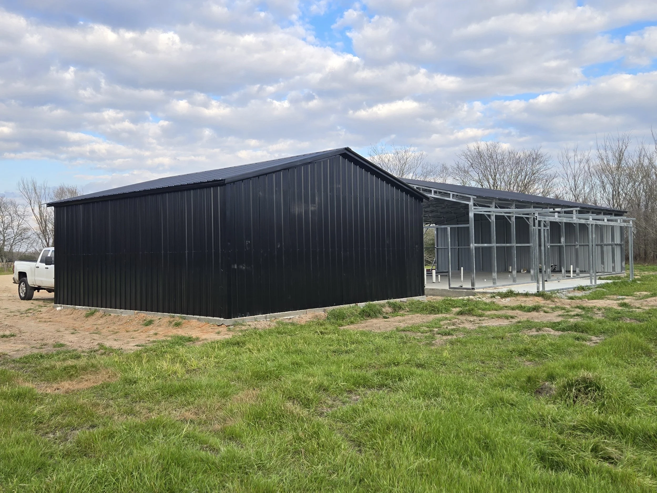 Large black metal building under construction with metal framing and a white pickup truck nearby, set on a grassy area with a cloudy sky overhead.