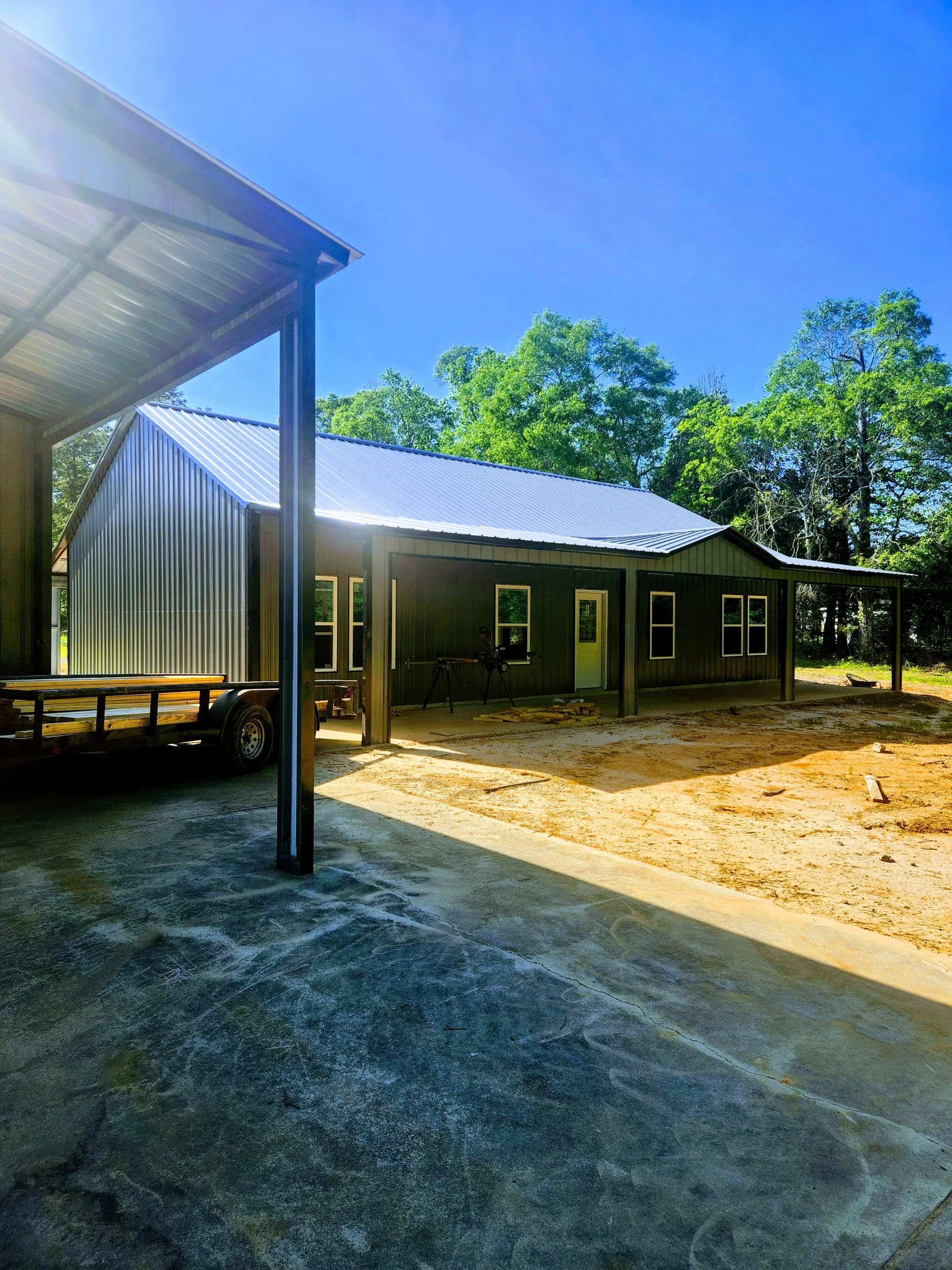 Construction site with a metal building, trees in the background, and a clear blue sky.