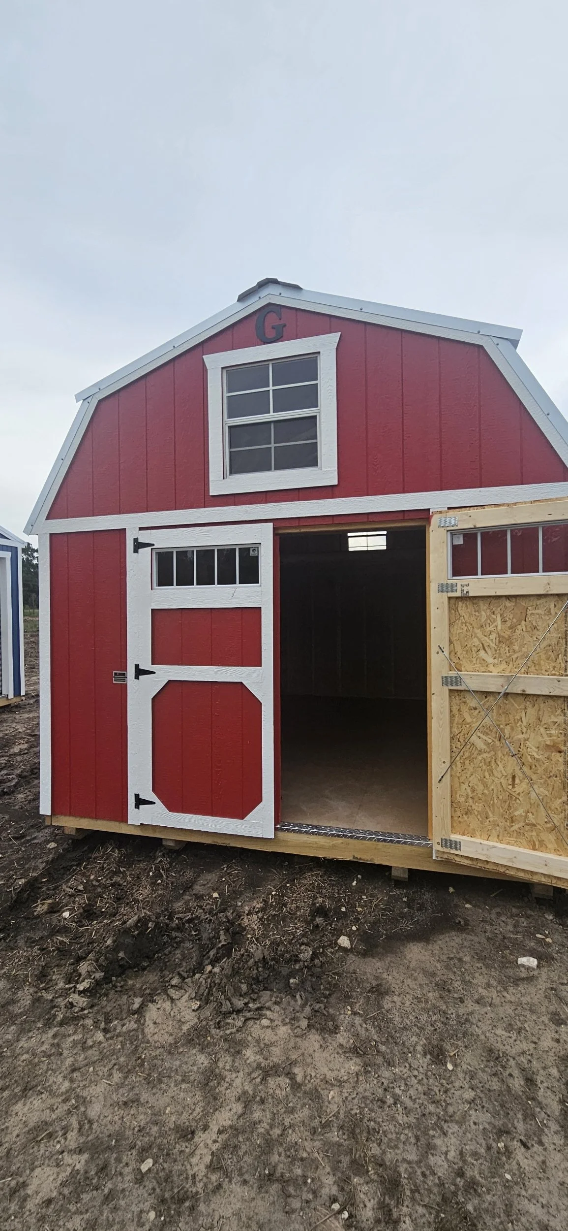 A red barn-style shed with a white-trimmed window and door, and an open doorway showing the interior, situated outdoors on dirt ground.