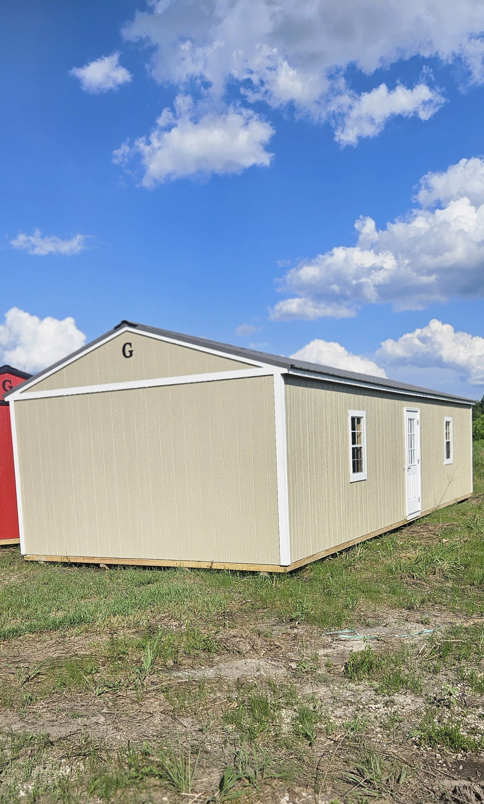 A beige portable building with white trim and a white door, set on grass, with a partly cloudy blue sky overhead.