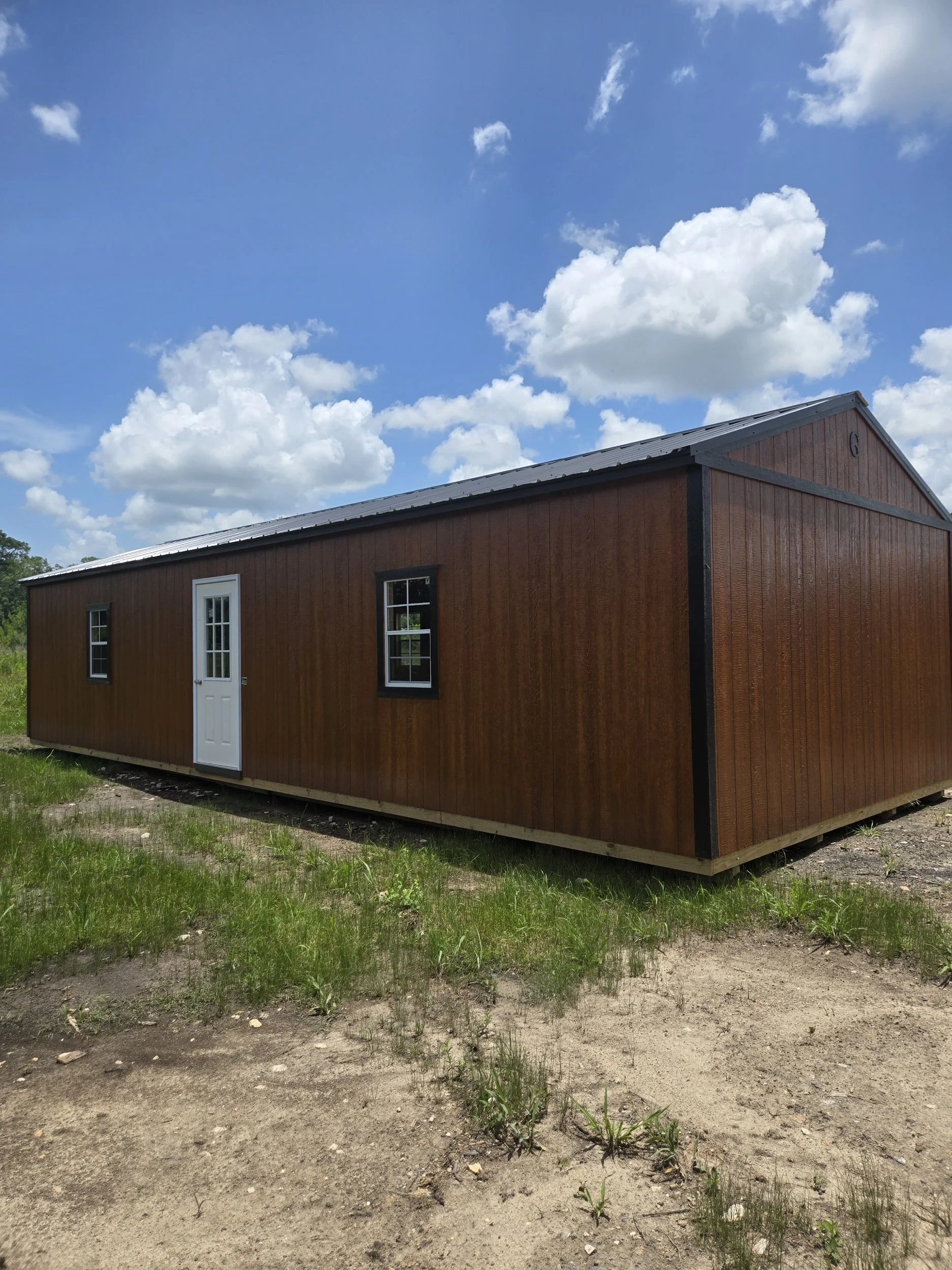 A long, rectangular wooden shed with a gray metal roof, white door, and two black-framed windows, situated on a patchy grassy area under a partly cloudy sky.