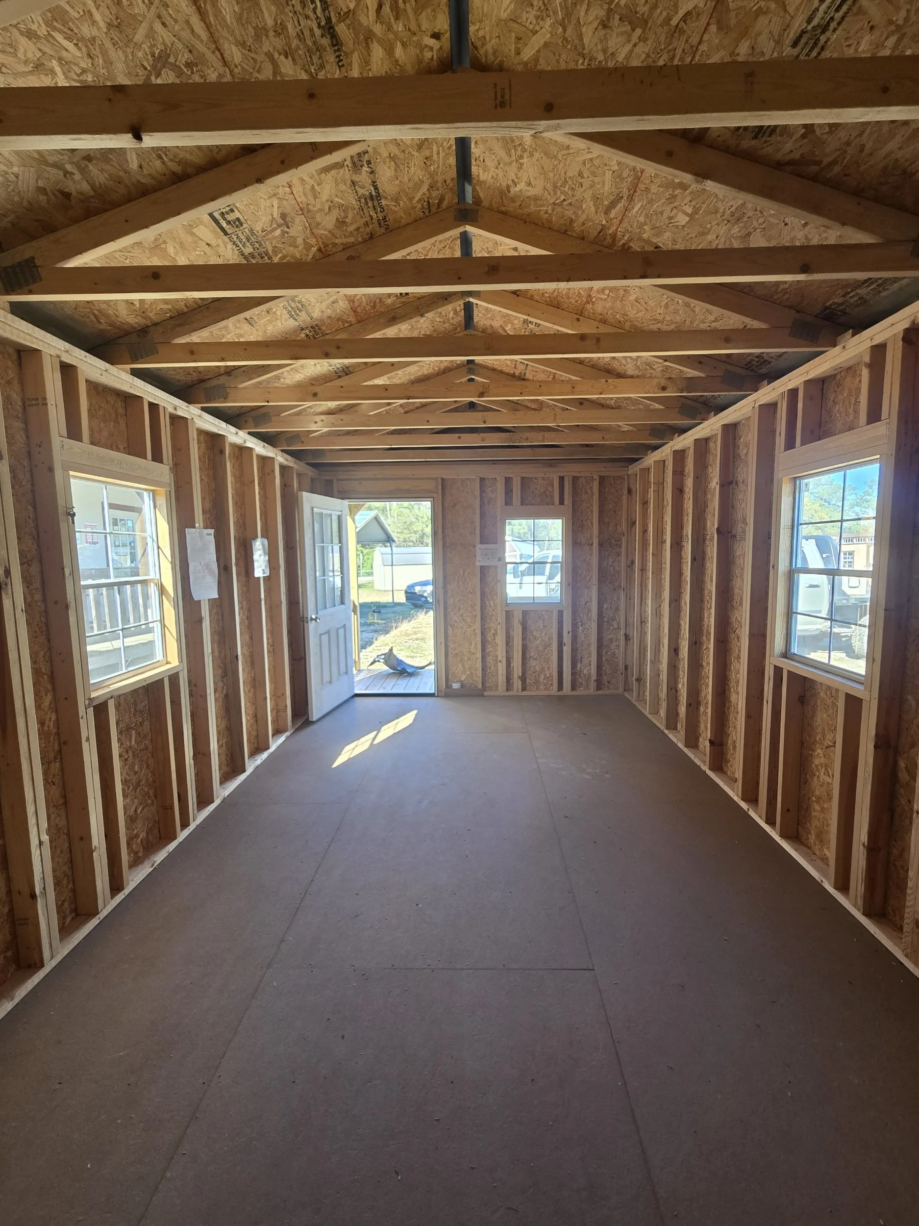 Interior of a small wooden structure under construction with exposed wall framing, roof trusses, three windows, a door, and a concrete floor.