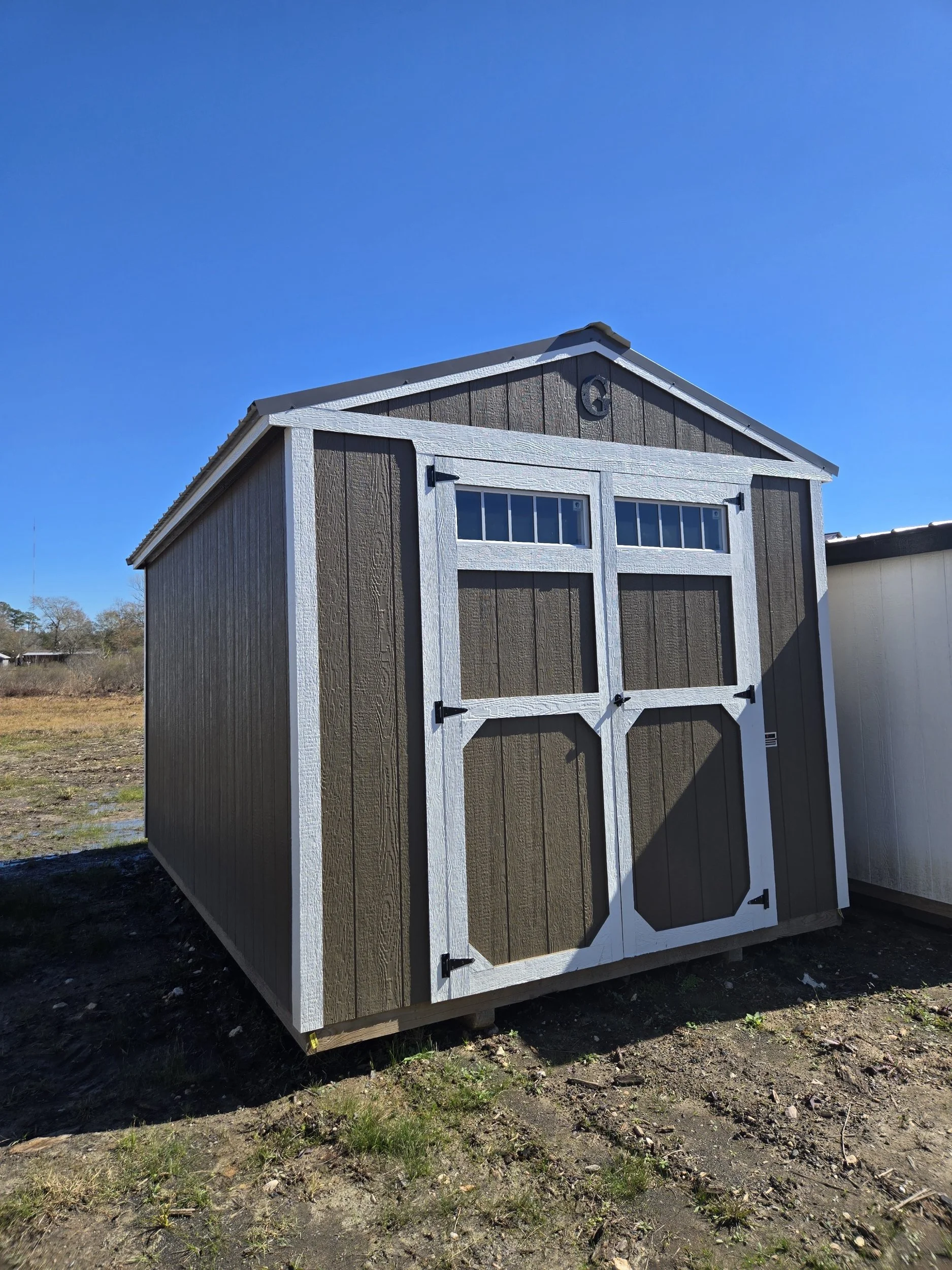 A small shed with brown vertical siding, white trim, and a double door with windows, set under a bright blue sky.