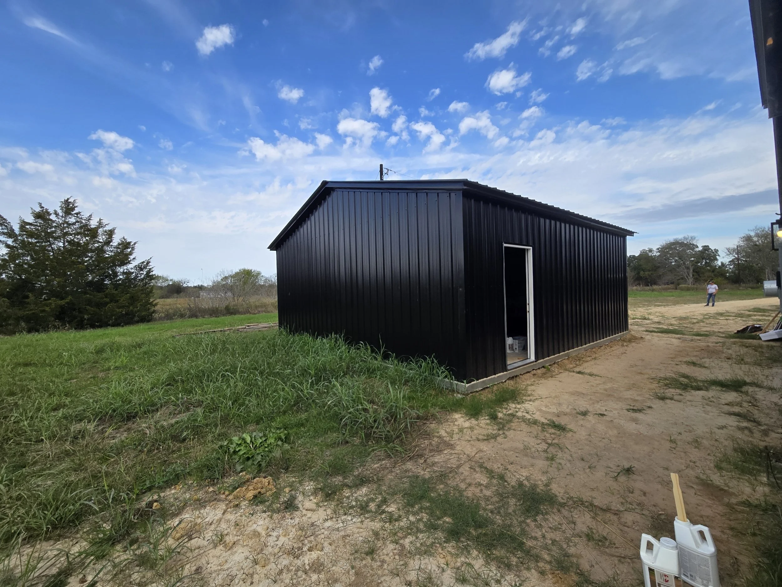 A black metal shed with an open door, situated on a dirt and grassy field under a partly cloudy blue sky, with two people visible in the distance.