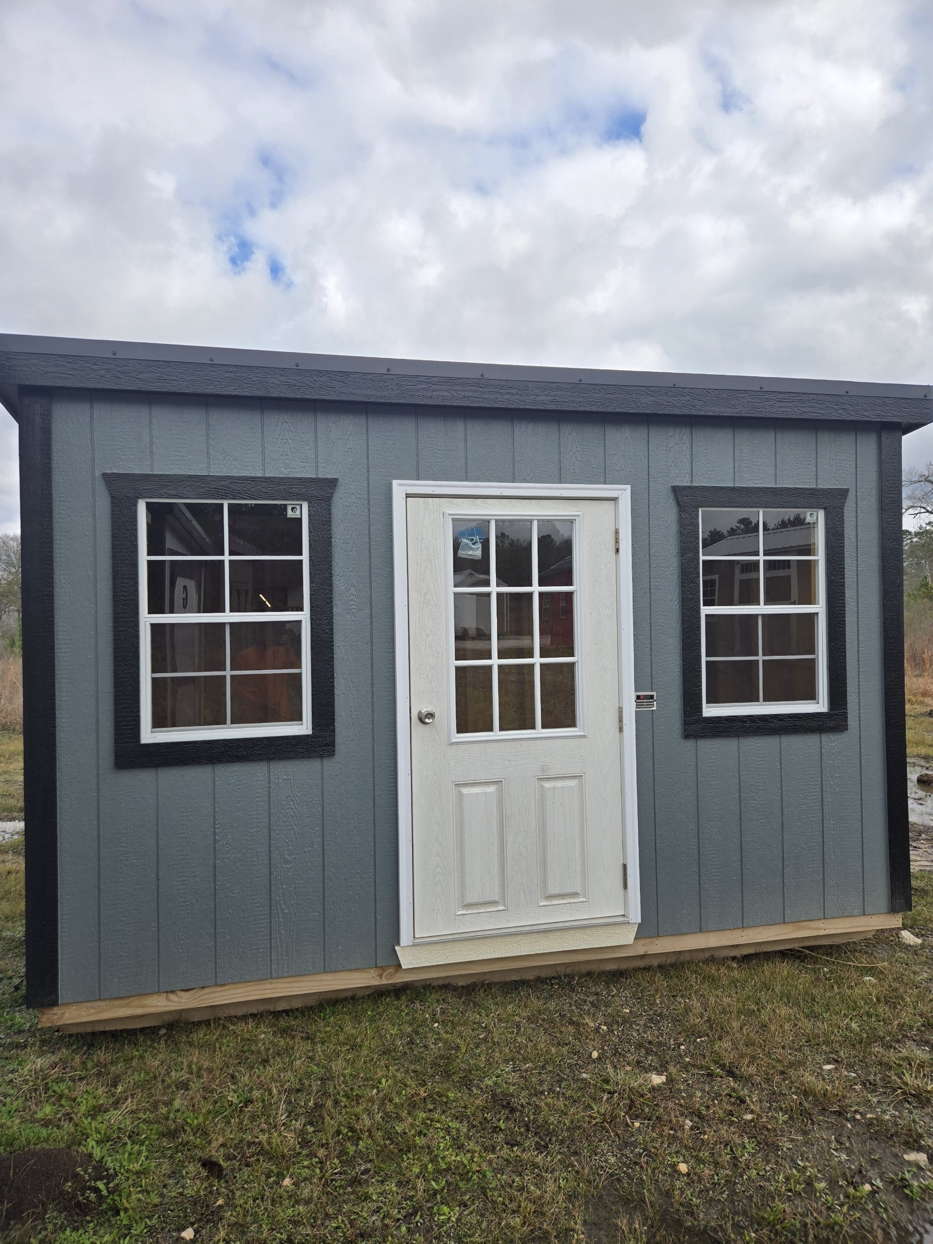 Small gray shed with black window trim, white door with glass panels, and a sloped roof, sitting on a grassy area under a partly cloudy sky.