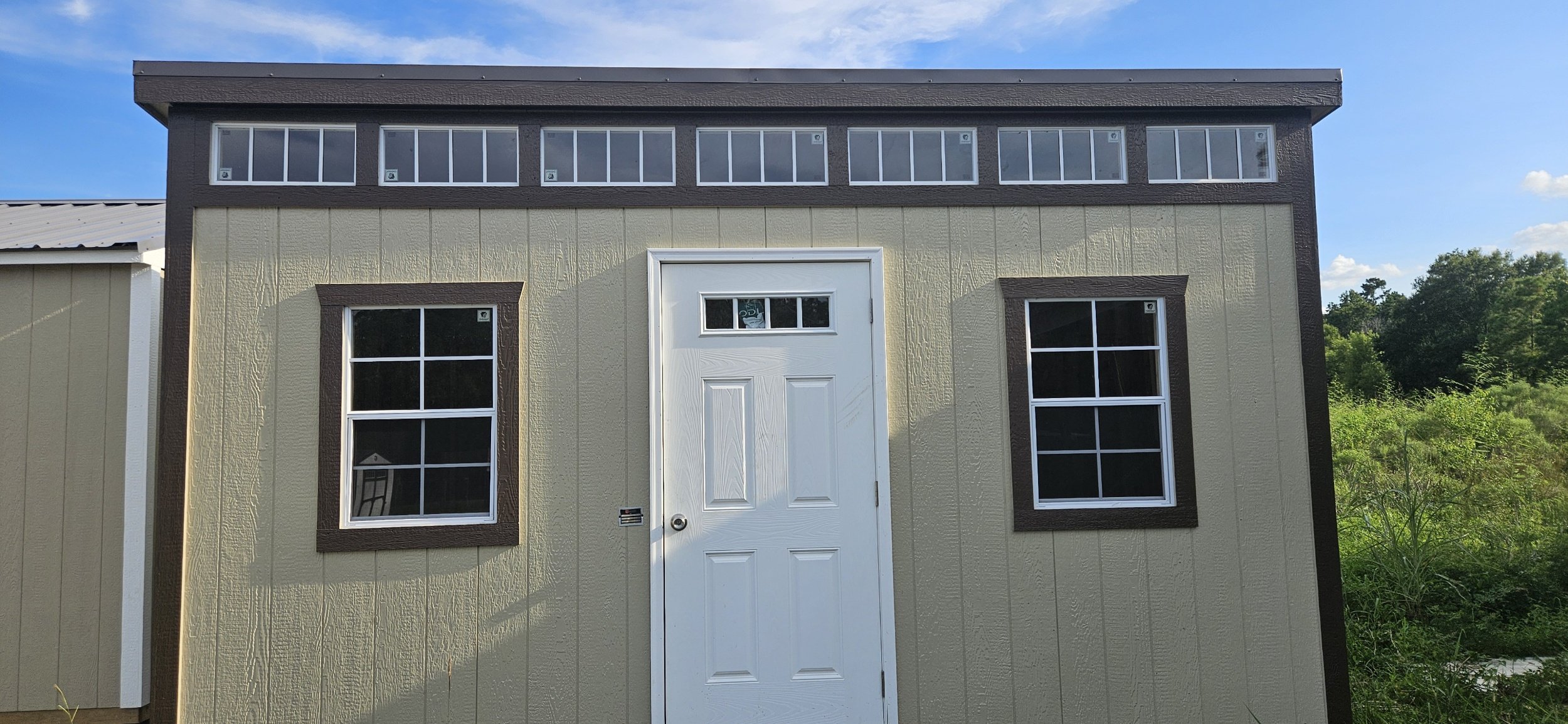 A small beige shed with brown trim, two windows on either side of the door, and a row of windows at the top, set against a blue sky with trees in the background.