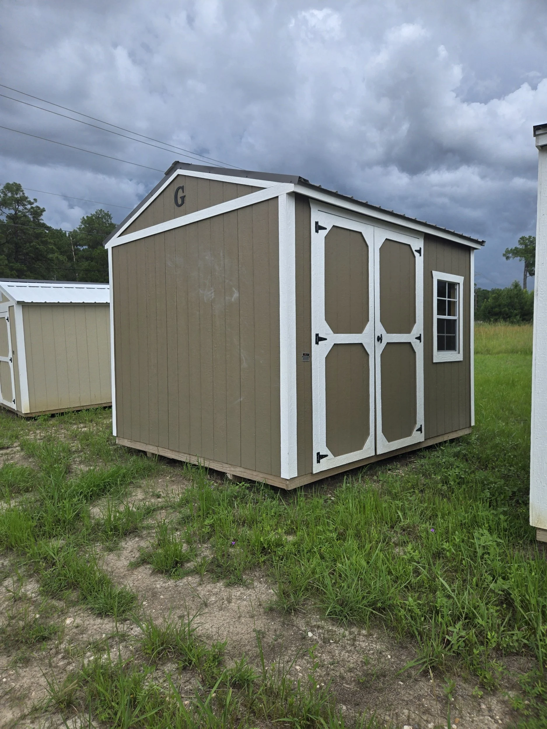 A small beige garden shed with white trim and double doors, situated on a grassy and sandy area under a cloudy sky.