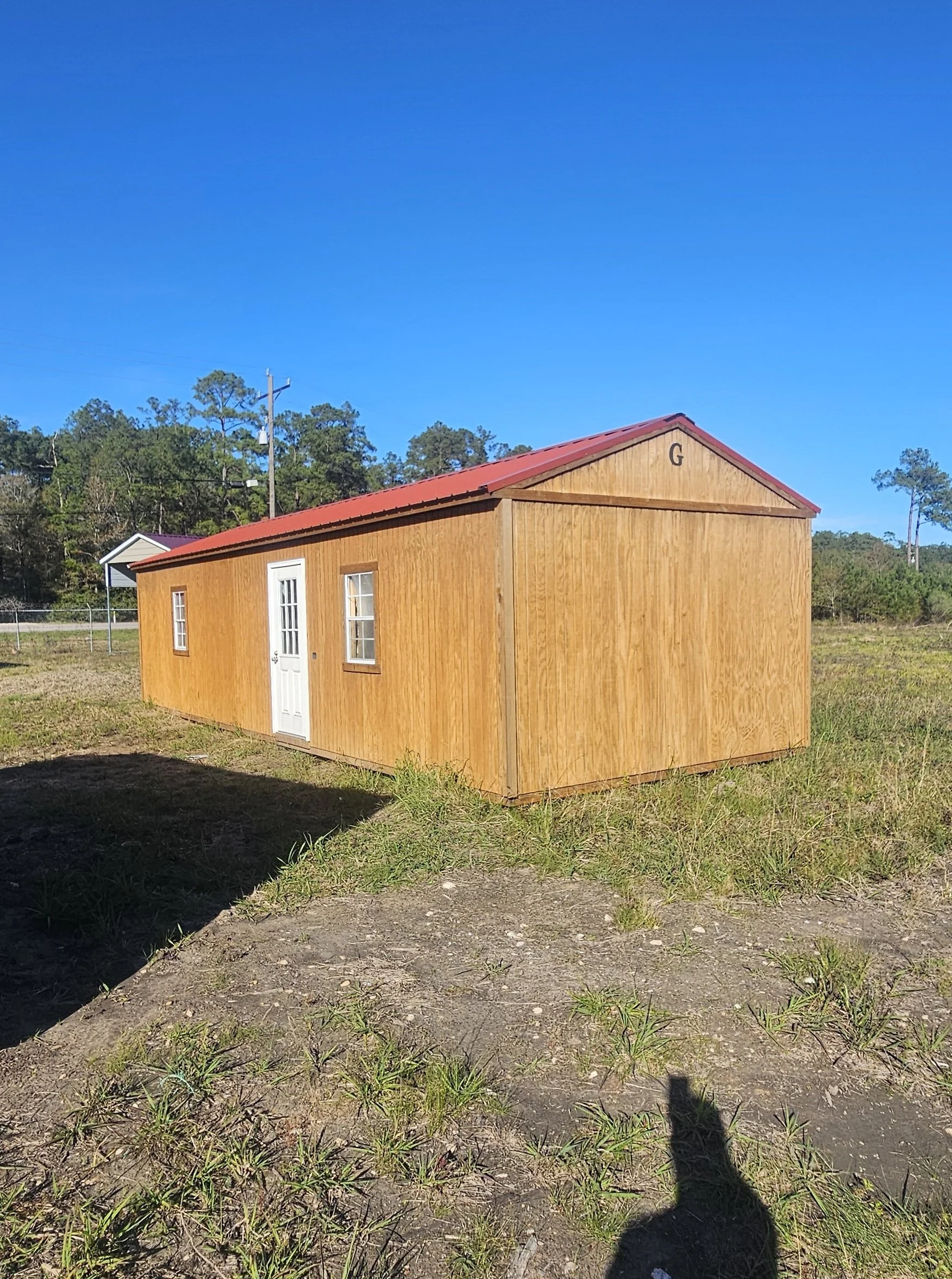 A small wooden shed with a red metal roof, white door, and two windows, situated on a grassy and dirt field with power lines and trees in the background under a clear blue sky.
