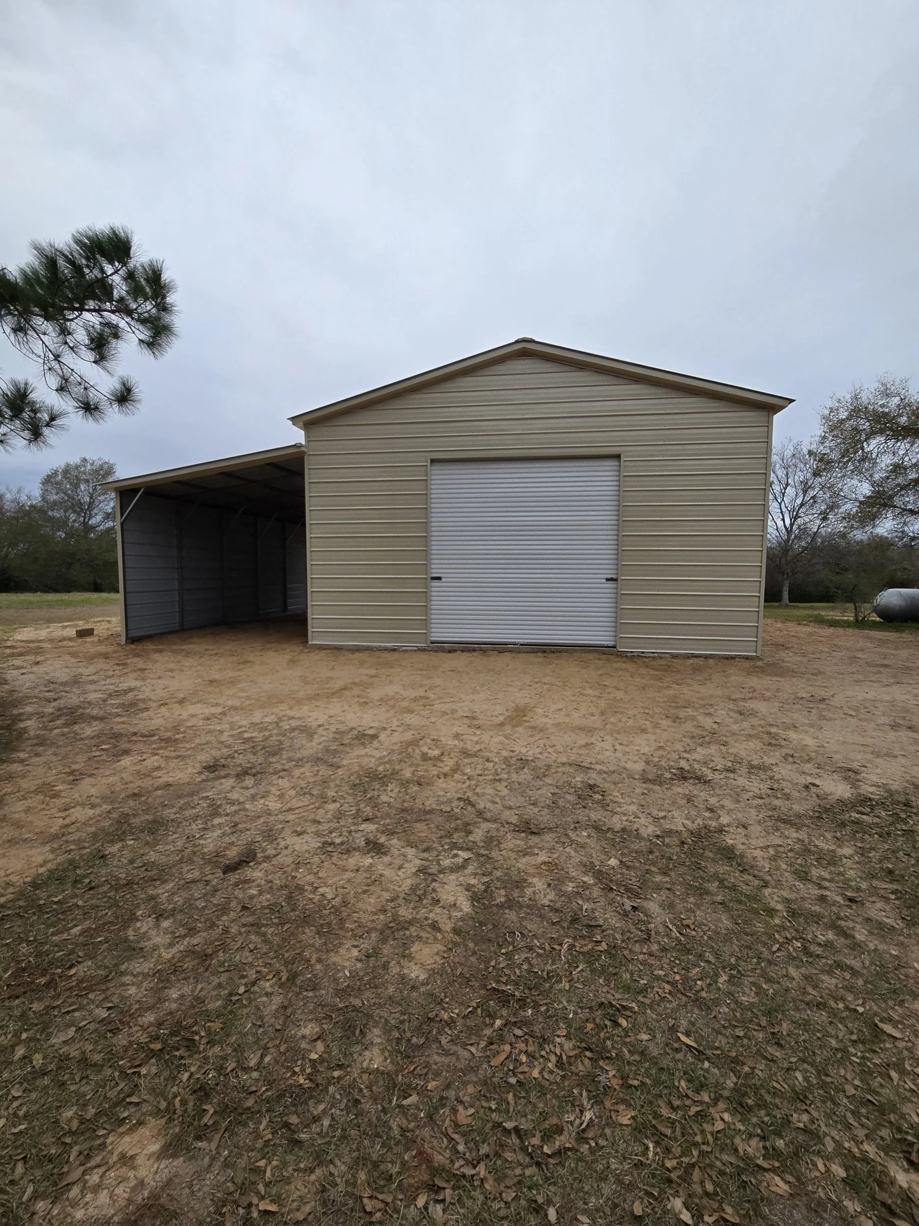 A beige and gray metal shed with a garage door in a rural outdoor setting, with dirt ground and some trees in the background.