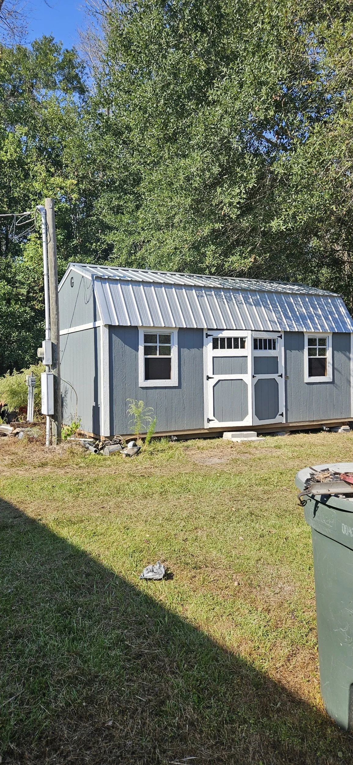 A small blue shed with white trim and black hardware, sitting on a grassy lawn next to a green trash can. The shed has two small windows and a double door with smaller window panes at the top.
